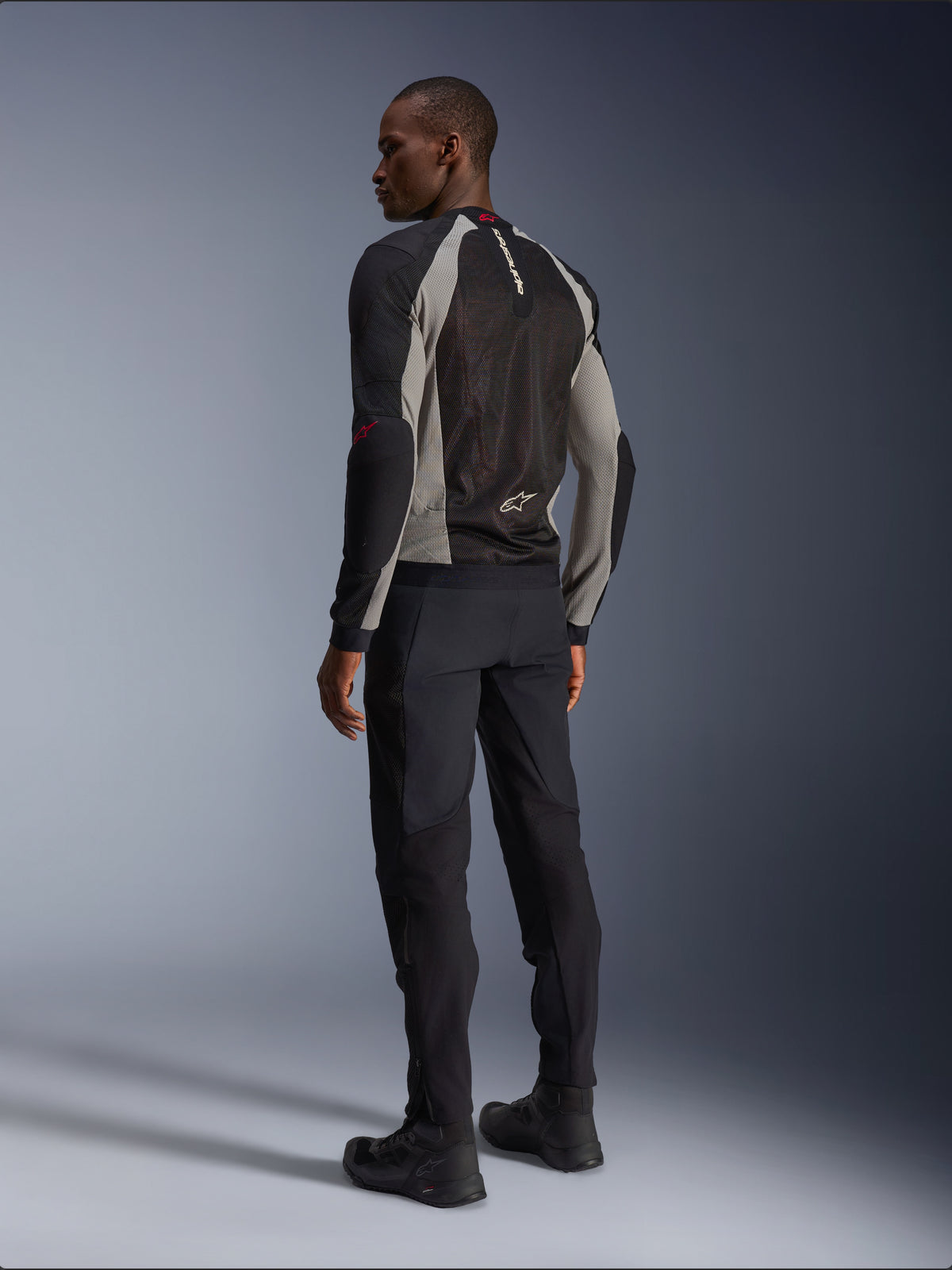 Model draagt de Alpinestars Troop-Air Jacket van achteren gezien, Black Ash Gray met felrode accenten, technische mesh-constructie voor ventilatie, gecombineerd met een zwarte motorbroek en motorlaarzen, urban contemporary stijl.