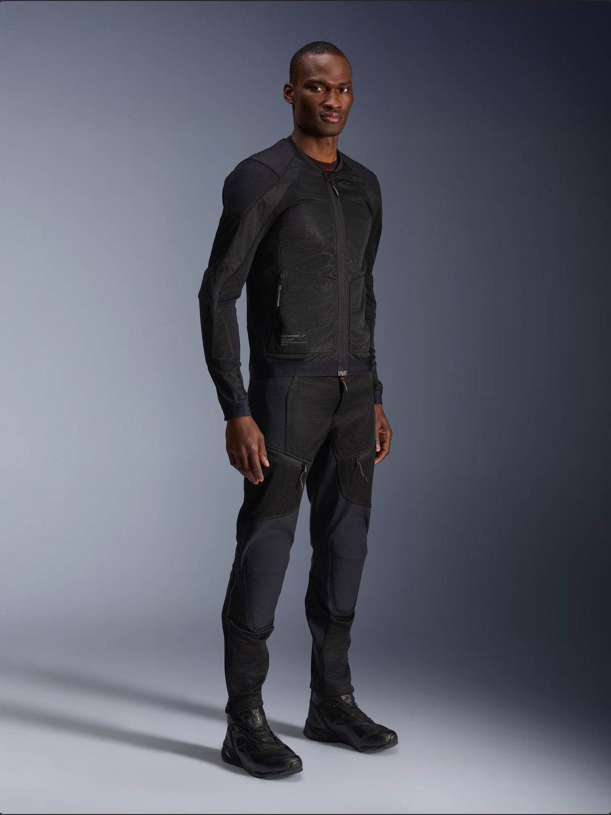 Alpinestars Troop-Air Jacke, Schwarz, technische Mesh-Motorradjacke getragen von einem Model mit passender schwarzer Hose und Stiefeln, leichte Mesh-Konstruktion, minimalistisches urbanes Design