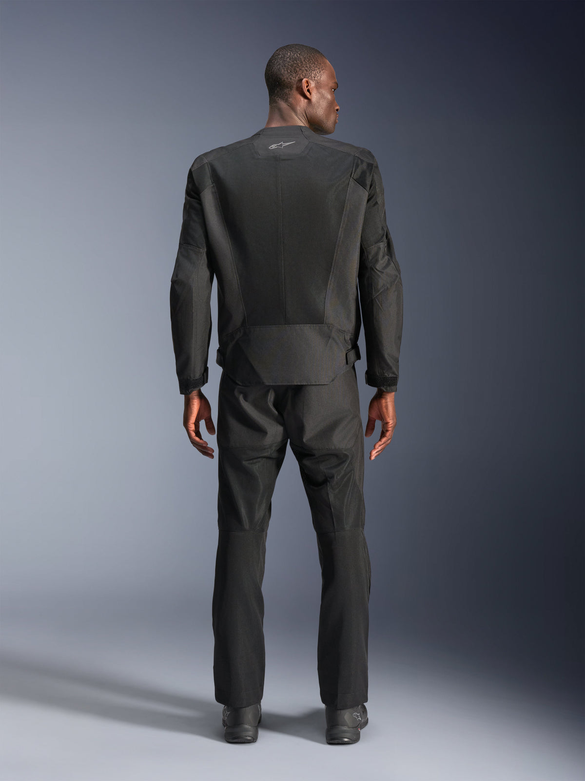 Rear view of a man wearing the Alpinestars C-1 Air Jacket in Black, urban motorcycle jacket with tech mesh panels, matching black textile pants and boots, clean aesthetic with reflective logo on the upper back.