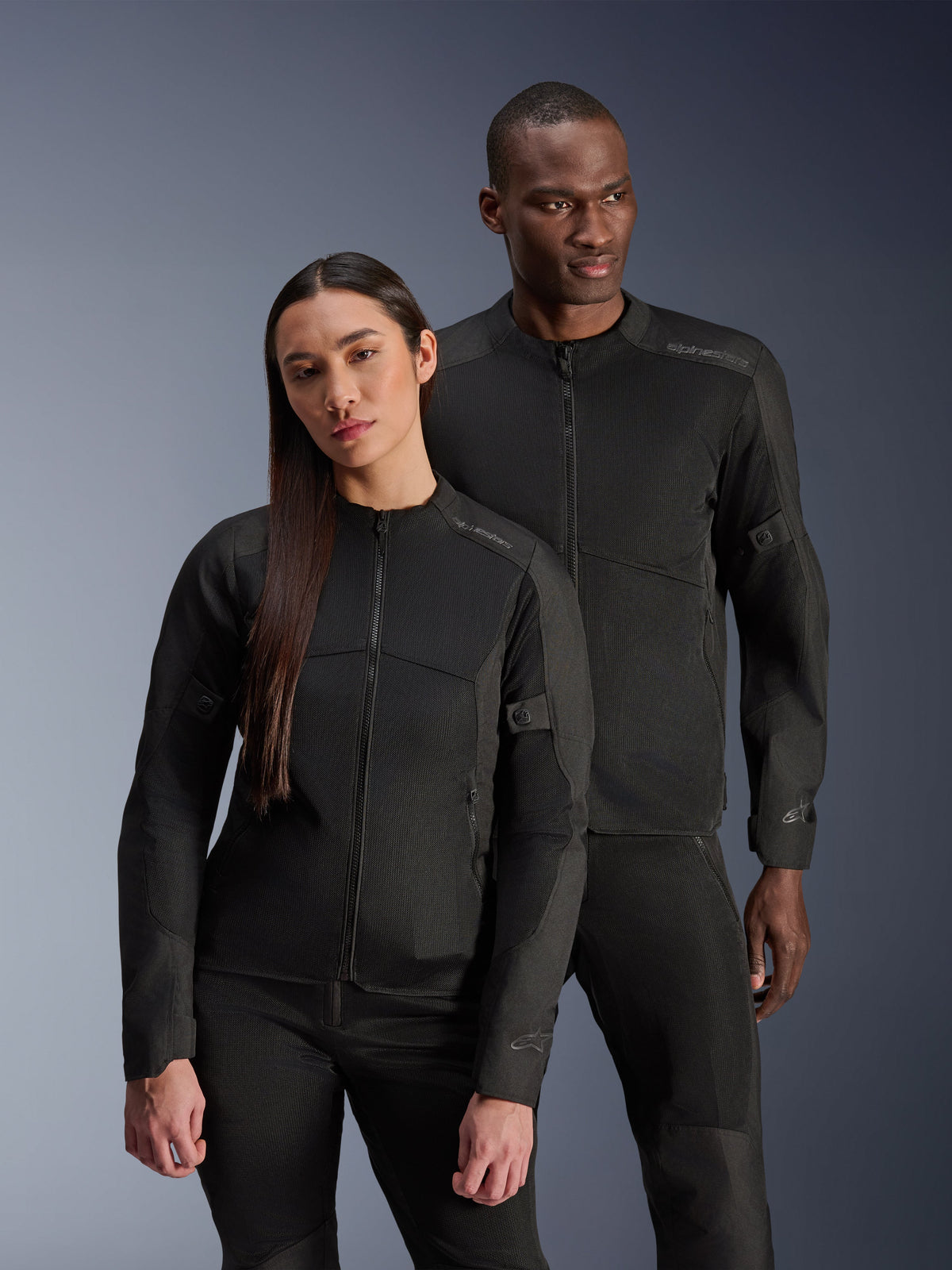 A man and a woman wearing the Alpinestars C-1 Air Jacket and Stella C-1 Air Jacket, matching black urban textile motorcycle jackets with mesh panels for ventilation, paired with black riding pants for a clean, contemporary look.
