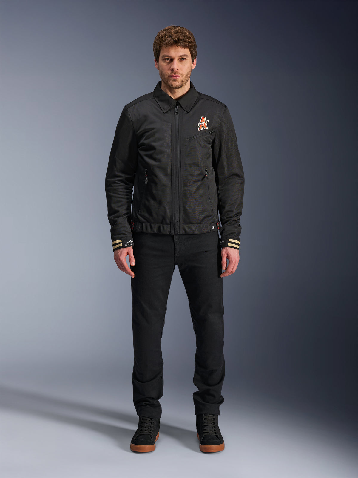 Mannelijk model draagt de zwarte Alpinestars Ignite Air Jacket, voorzien van een klassieke overhemdkraag, een volledige ritssluiting aan de voorzijde en een oranje logo op de borst. Gecombineerd met een zwarte broek en zwarte hoge sneakers voor een eigentijdse urban motorlook.
