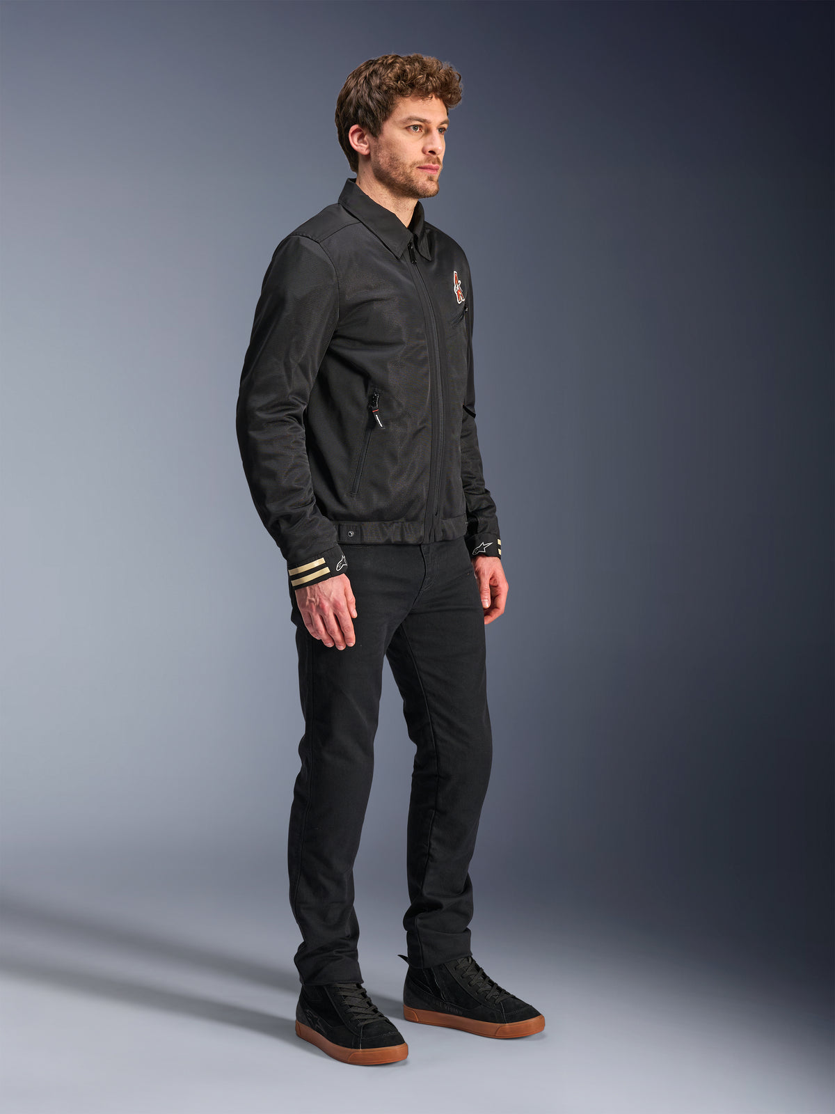 Alpinestars Ignite Air Jas, urban motorjas, zwart, gedragen door een model met zwarte broek en zwarte sneakers, voorzien van een traditionele overhemdkraag, club-geïnspireerde graphics op de borst en manchetten, strak eigentijds design met mesh en satijnen afwerking