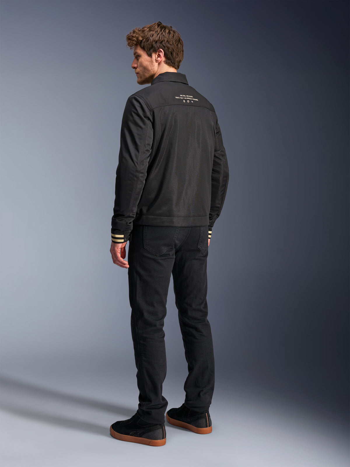 Alpinestars Ignite Air Jas, zwart motorjack, achteraanzicht met witte grafische tekst op de bovenrug en beige gestreepte manchetten, Harrington-stijl silhouet met mesh body, gecombineerd met een zwarte broek en zwarte sneakers