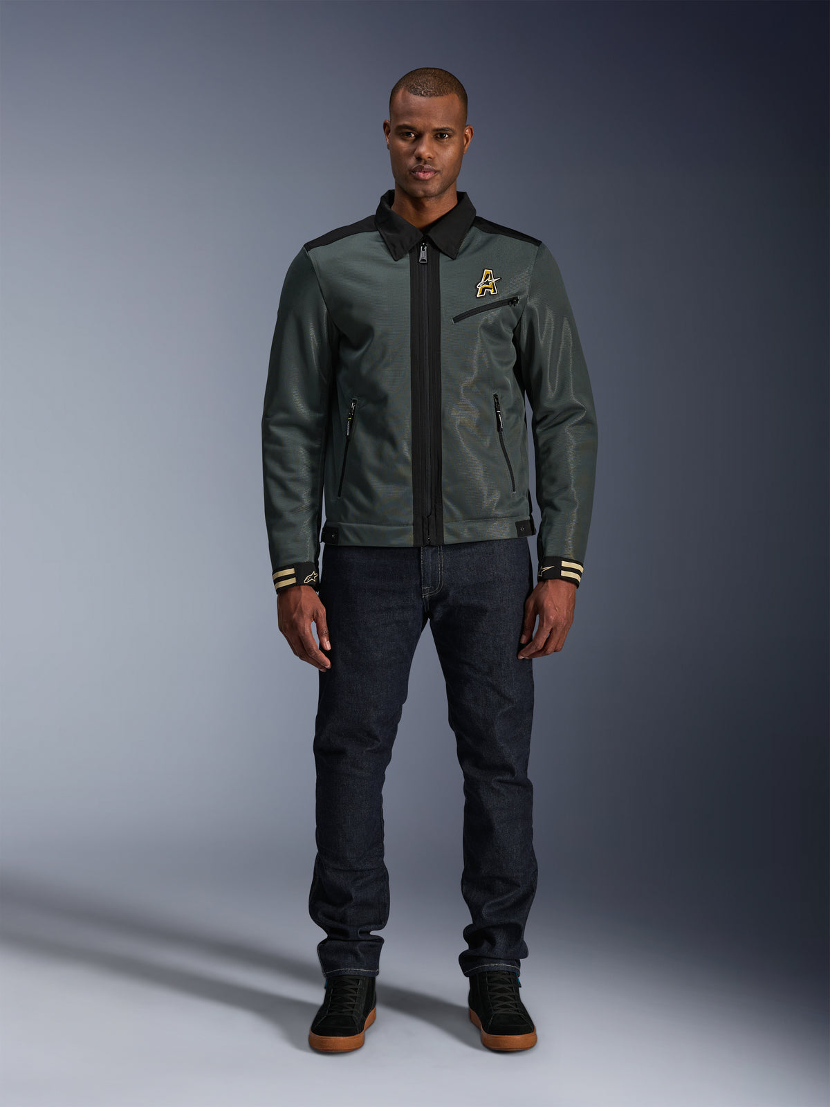 Alpinestars Ignite Air Jas in Urban Gray Black Blue, Harrington-stijl motorjas met overhemdkraag en mesh-constructie, gedragen door een man met donkere denim jeans en zwarte sneakers, urban streetwear voor motorrijders.