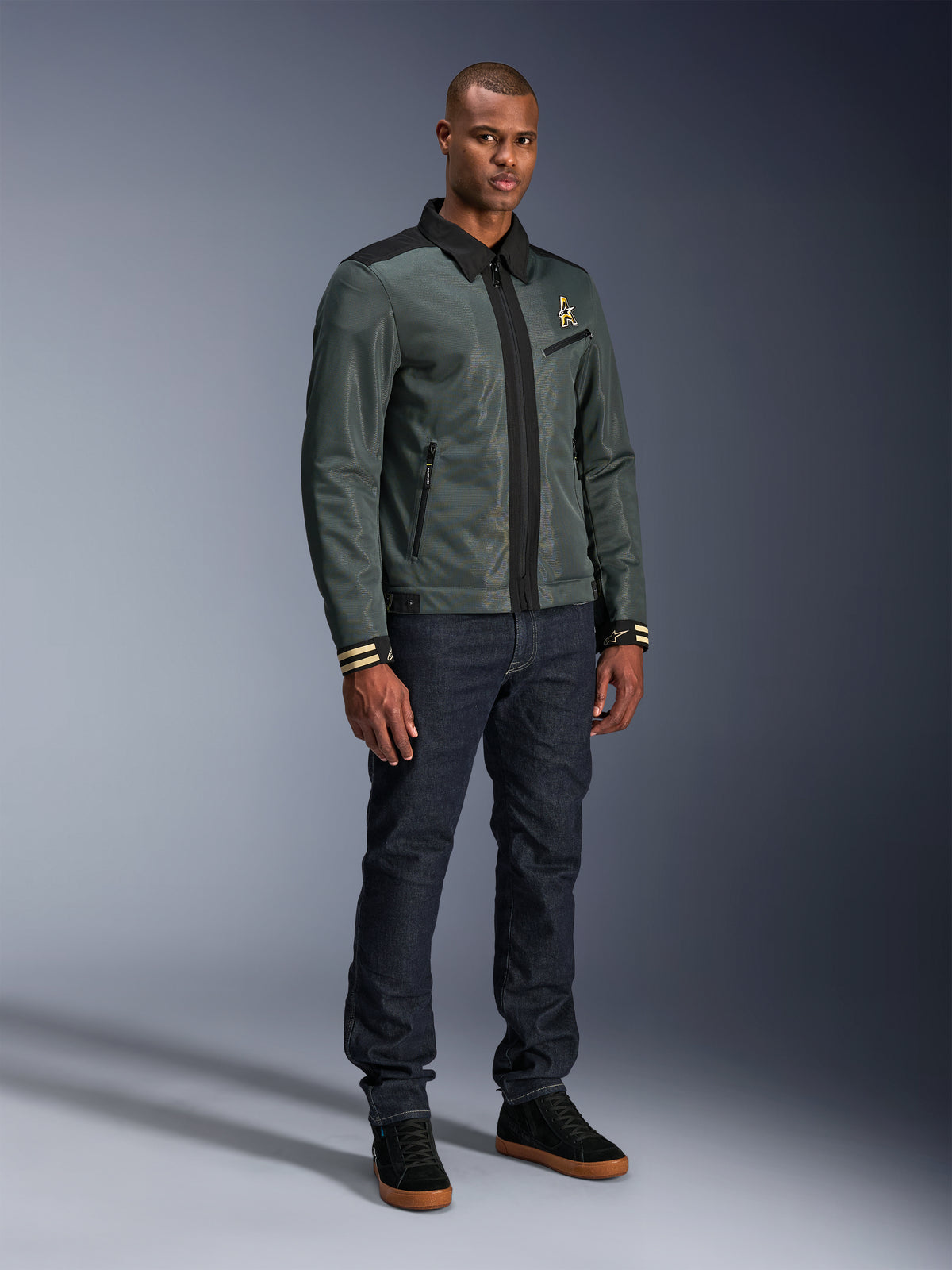 Alpinestars Ignite Air Jacket, Urban Gray Black Blue, urban motorjas, gedragen door een model met donkere denim jeans en zwarte sneakers, voorzien van een mesh body, traditionele overhemdkraag, Harrington-geïnspireerd silhouet en gouden Alpinestars branding.