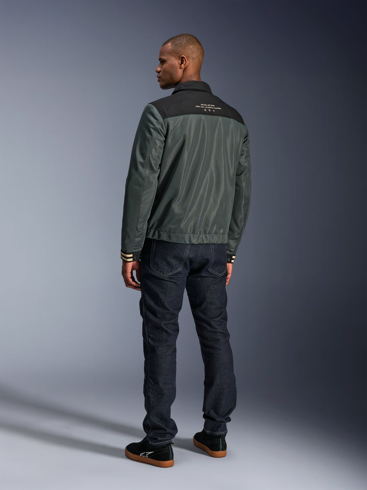Alpinestars Ignite Air Jas in Urban Gray Black Blue, achteraanzicht op model, met een mesh body, een zwart schouderstuk met witte tekst en gestreepte manchetten, gecombineerd met een donkere denim jeans en zwarte sneakers, urban lifestyle motorkleding