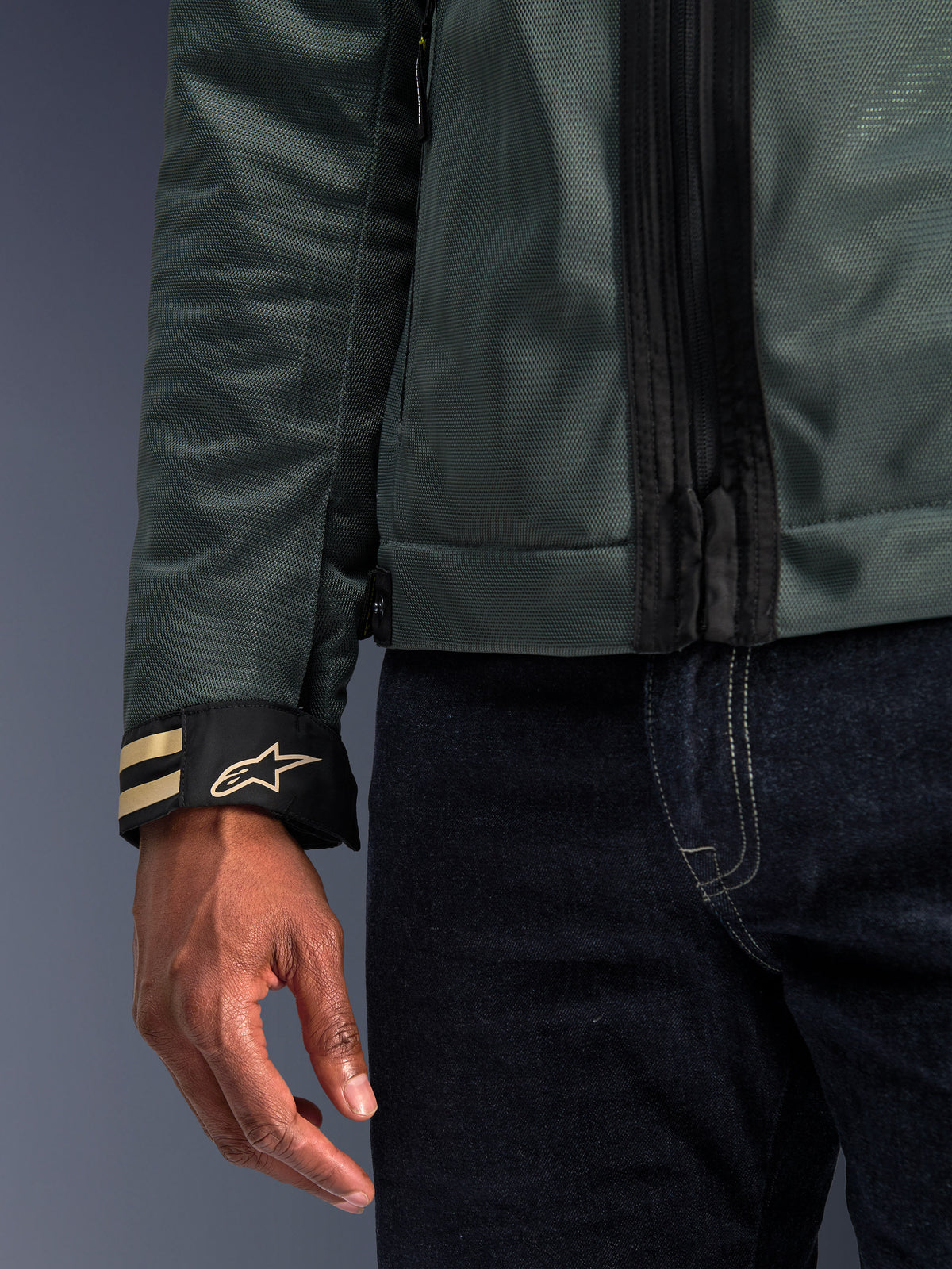 Alpinestars Ignite Air Jas in Urban Gray, close-up van mouw met mesh-stof en zwarte manchet met gouden strepen en ster-logo, gedragen met donkerblauwe denim jeans, eigentijdse stedelijke motoruitrusting.