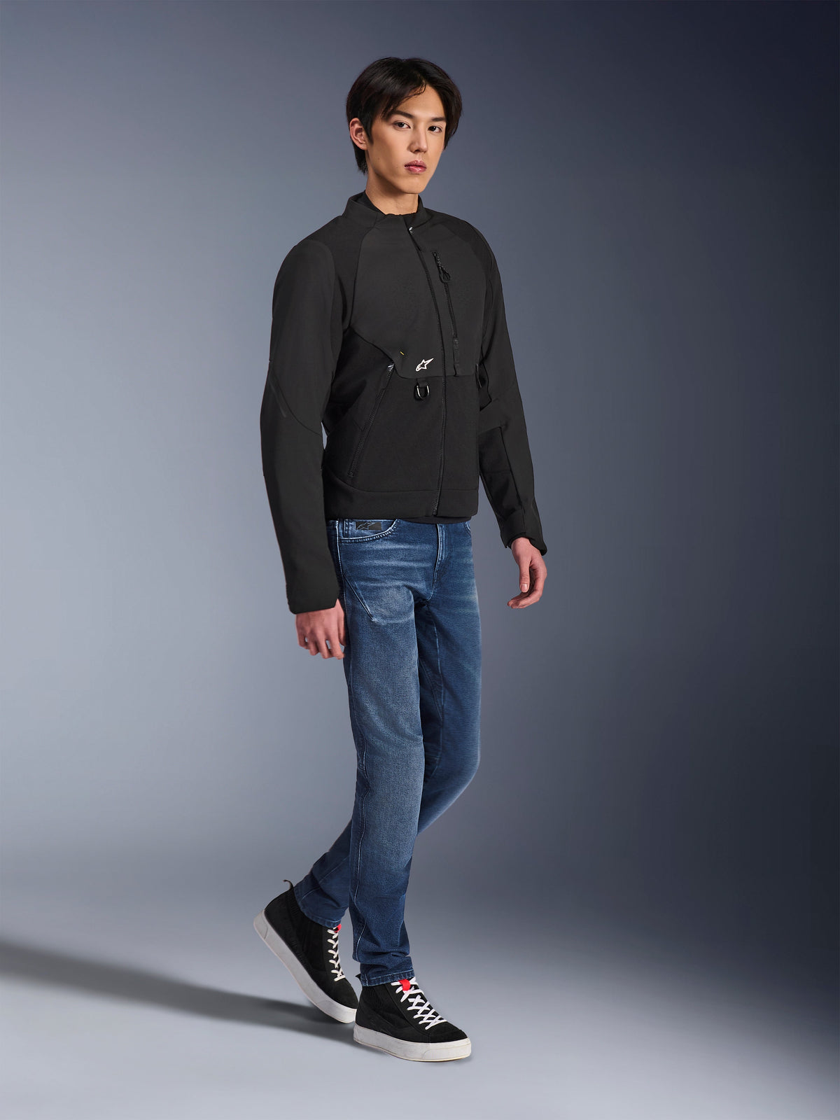 Veste Alpinestars Provoke, veste de moto urbaine, noire, portée par un mannequin homme avec un jean bleu et des baskets noires, design en textile technique avec petit logo Alpinestars, photo lifestyle en pied pour la conduite urbaine contemporaine