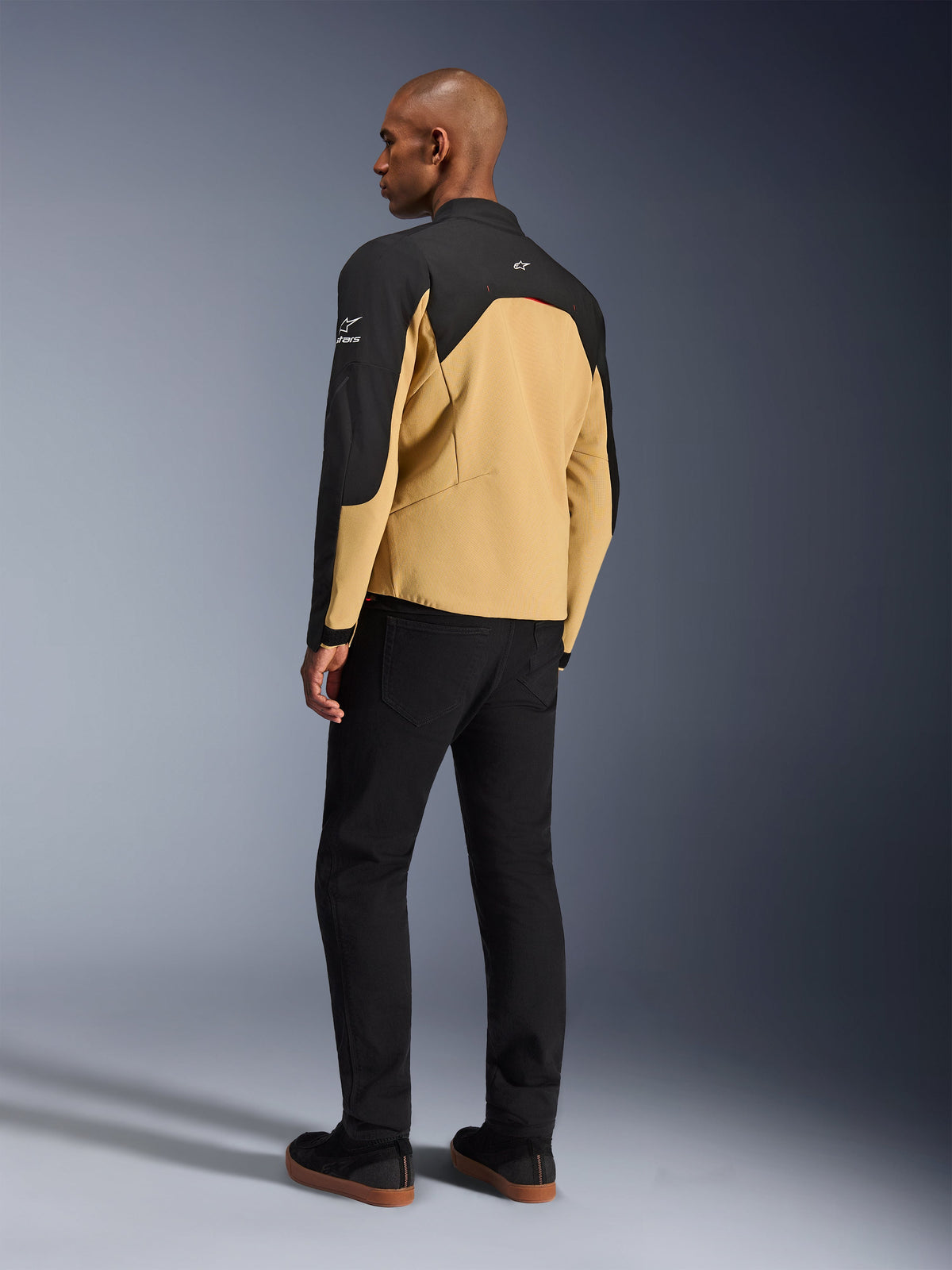 Alpinestars Provoke Jacke, urbane Motorradjacke in Black Camel, Dreiviertel-Rückansicht, camel-farbener Rumpf mit schwarzen Schulter- und Ärmeleinsätzen, weiße Alpinestars-Logos, getragen mit schwarzer Hose, moderne technische Fahrausrüstung