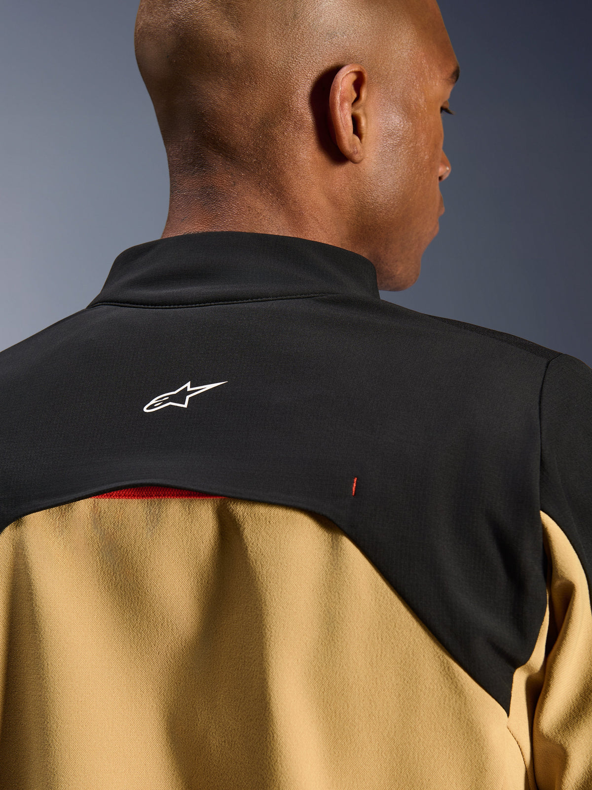 Alpinestars Provoke Jacke, Urbane Motorradjacke, Schwarz und Camel, Nahaufnahme des oberen Rücken- und Kragenbereichs mit weißem Astars-Logo auf schwarzer Rückenpasse und hellbraunen Paneelen, zeitgenössisches, von Outdoor-Bekleidung inspiriertes technisches Design