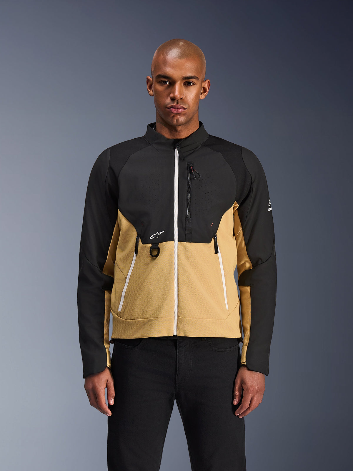 Alpinestars Provoke Jacke, Urbane Motorradjacke, Schwarz und Camel, Vorderansicht an einem männlichen Model, zweifarbiges Design mit weißen Reißverschlüssen, Brusttasche mit Reißverschluss und D-Ring, kleines weißes Alpinestars-Logo, moderne urbane Motorradbekleidung