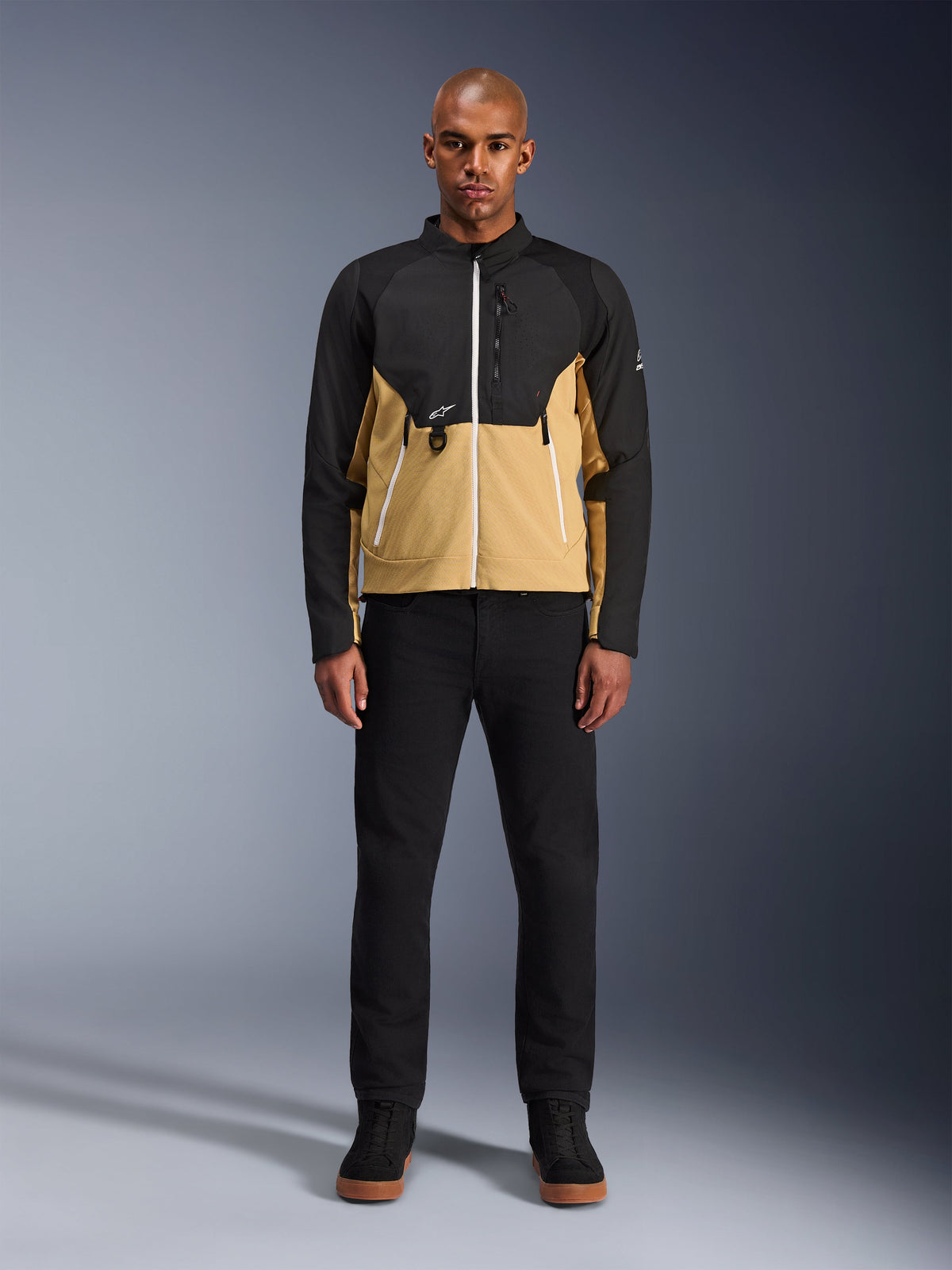 Alpinestars Provoke Jacke in Black Camel, urbane Motorradjacke mit zweifarbigem Design in Schwarz und Camel, getragen von einem Model mit schwarzer Hose und schwarzen Schuhen, moderne Silhouette mit weißen Reißverschlüssen und technischer Softshell-Konstruktion.