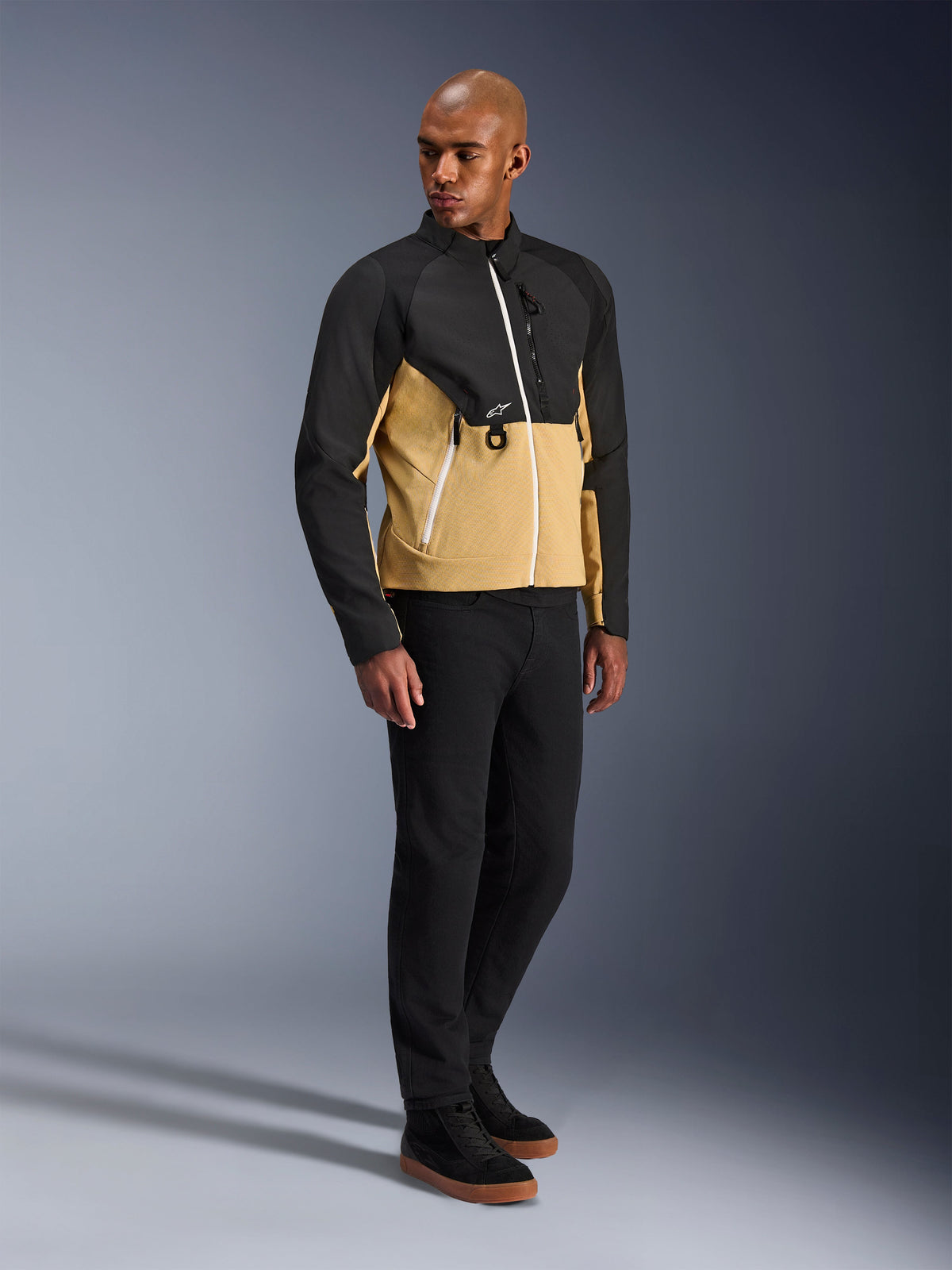 Alpinestars Provoke Jacke, urbane Motorradjacke, Black Camel, getragen von einem Model mit schwarzer Hose und Sneakern, zweifarbiges Textildesign mit schwarzem Ober- und camel-farbenem Unterkörper, weiße Reißverschlussdetails, moderne urbane Fahrerausrüstung