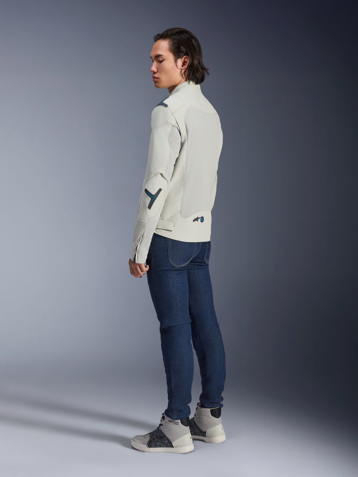 Alpinestars Circal-1 Air Jacke, urbane Motorradjacke, Metal Gray mit Methyl Blue Akzenten, von einem Model in der Dreiviertel-Rückansicht getragen, mit dunkelblauen Jeans und grauen High-Top-Sneakern, mit Mesh-Einsätzen und minimalistischem Design