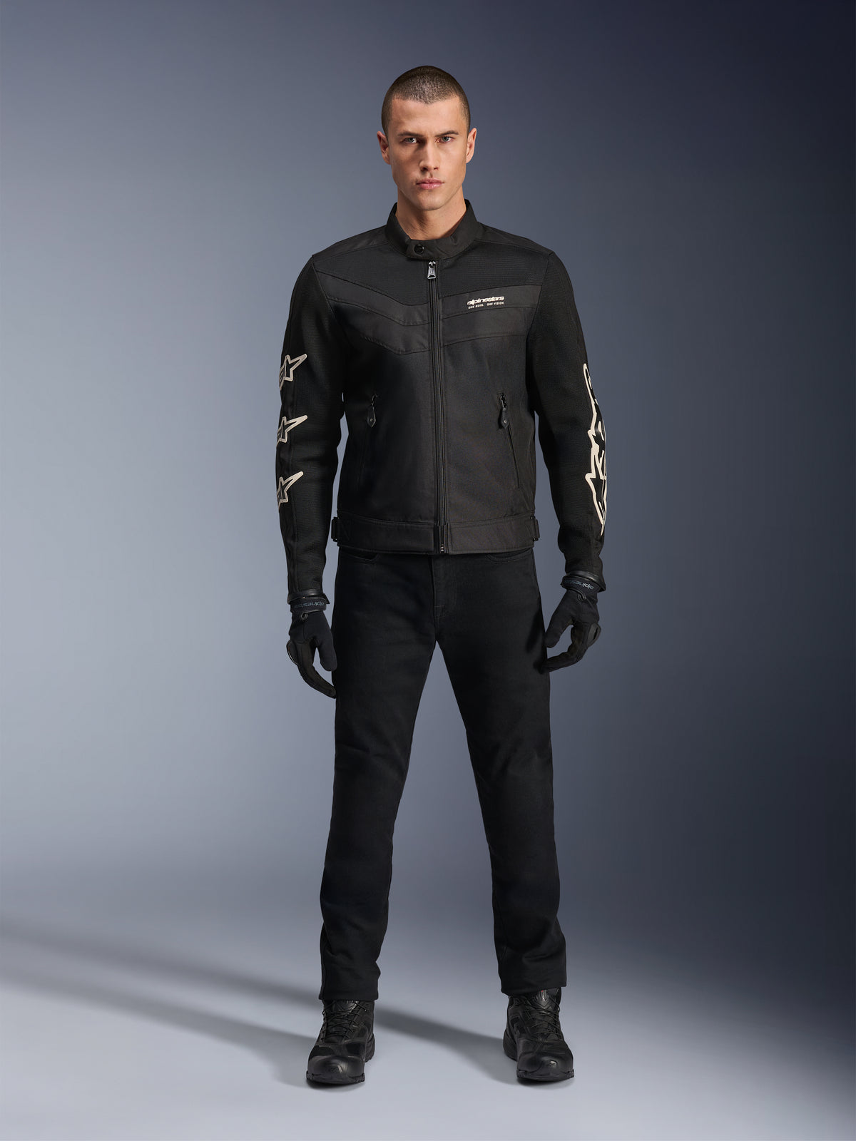 Männliches Model trägt die Alpinestars T-Dyno Air Jacke, schwarze, leichte Textil-Motorradjacke mit atmungsaktivem 3D-Mesh, weißen Stern-Logos auf den Ärmeln, kombiniert mit schwarzer Hose, Handschuhen und Stiefeln, Vorderansicht stehend