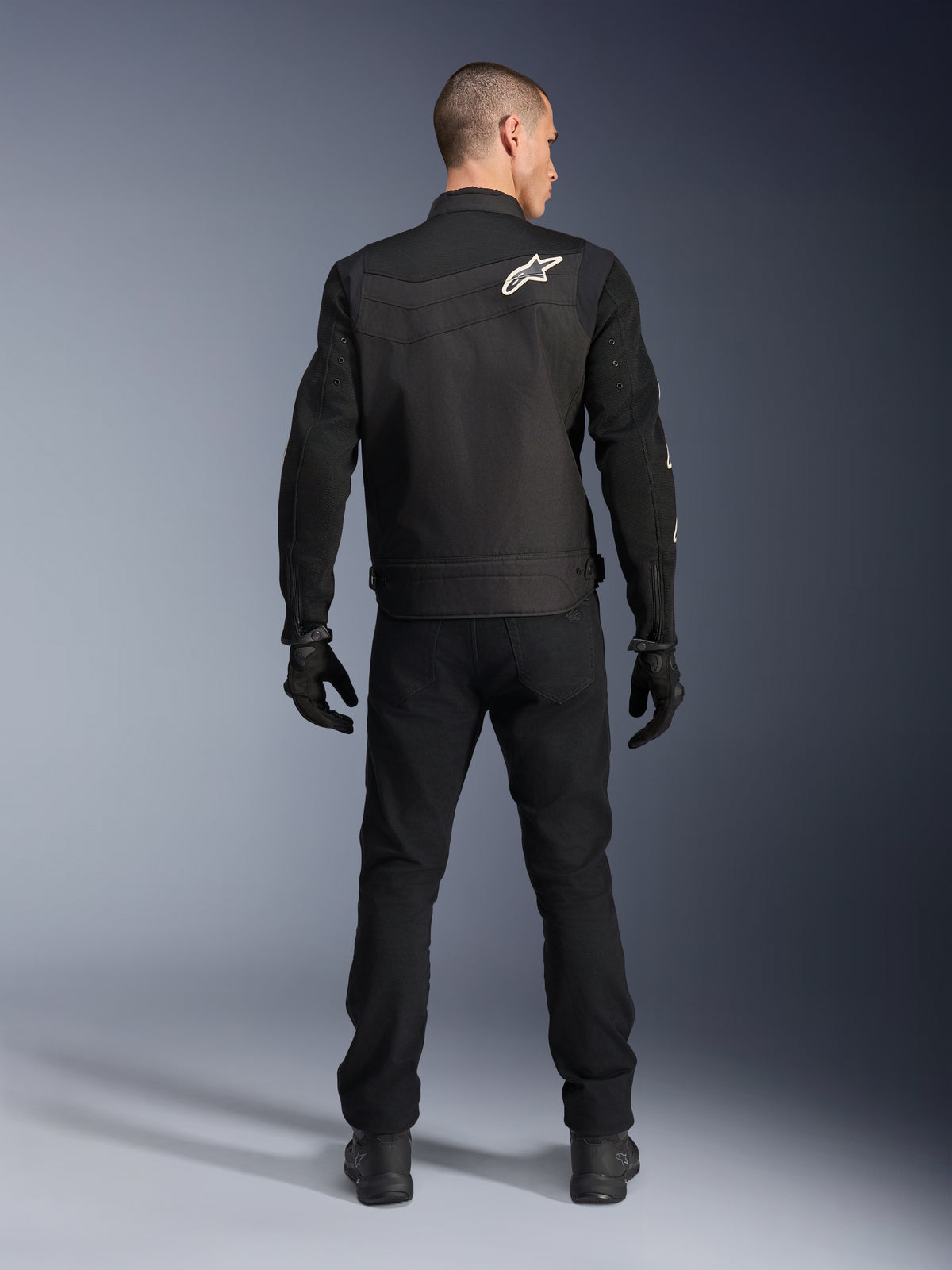Alpinestars T-Dyno Air Jacke, Schwarz, Rückansicht am Modell, Textil-Motorradjacke mit 3D-Mesh-Einsätzen und einem weißen Stern-Logo auf der Schulter, getragen mit schwarzer Hose und Handschuhen, modernes urbanes Fahroutfit