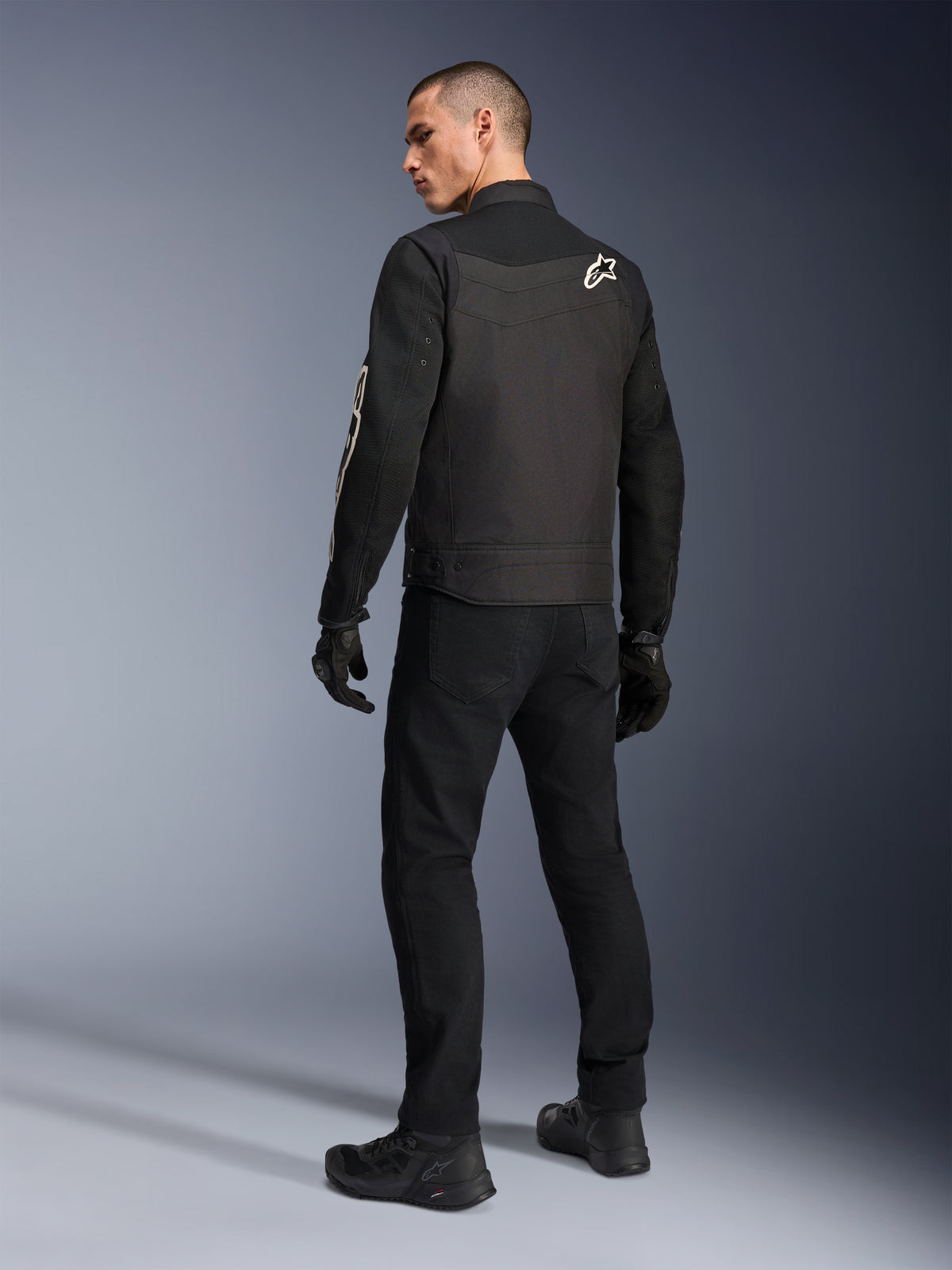 Alpinestars T-Dyno Air Jacke, Schwarz, Rückansicht eines Models in einer atmungsaktiven Textil-Motorradjacke, mit weißem Alpinestars-Logo auf der Schulter, kombiniert mit schwarzer Hose und Handschuhen, urbaner Fahrstil
