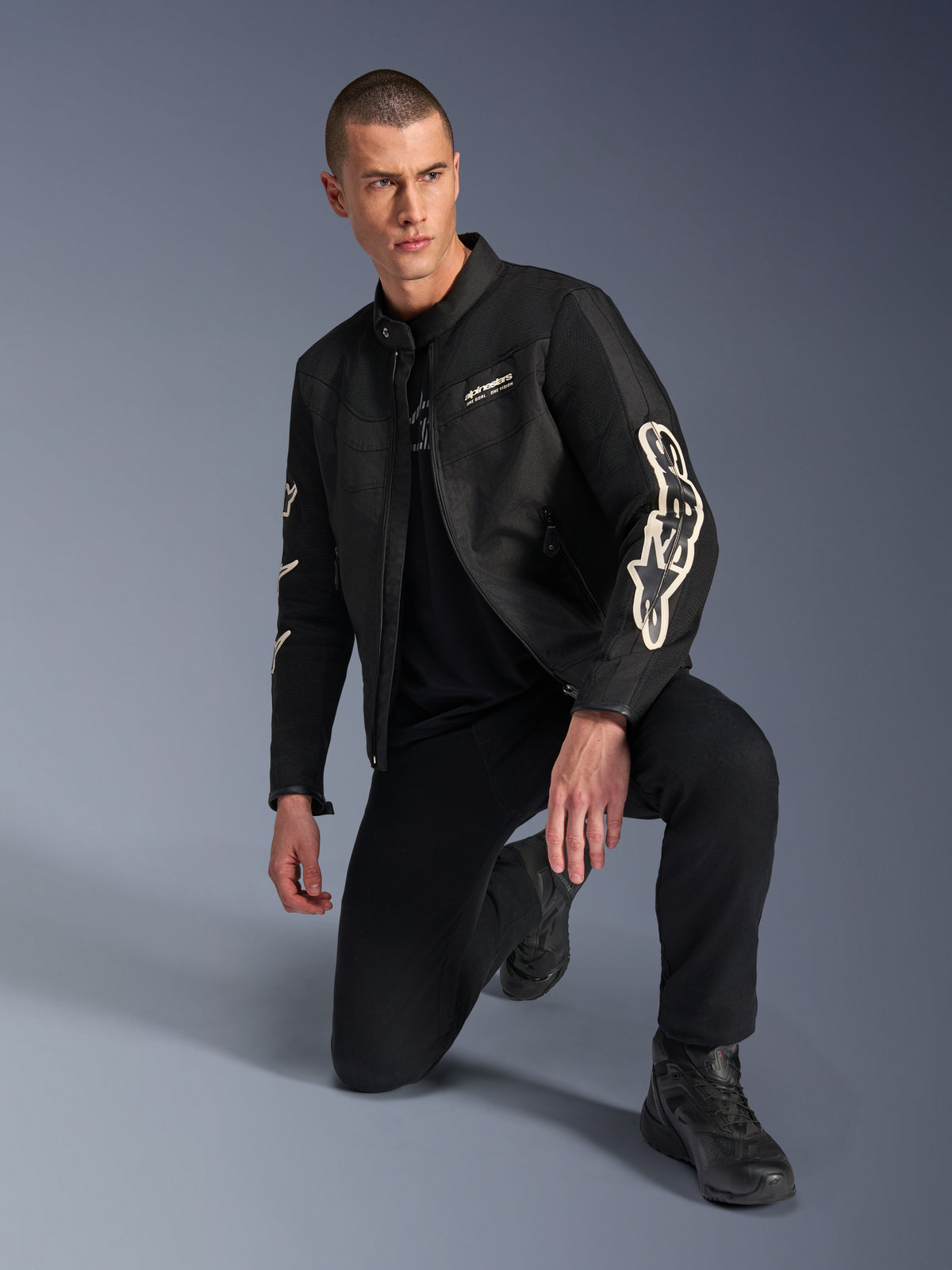 Männliches Model in Alpinestars T-Dyno Air Jacke, schwarze Textil-Motorradjacke mit großem weißen Branding auf den Ärmeln, kombiniert mit schwarzer Hose und Motorradstiefeln, modernes Vintage-inspiriertes Design mit 3D-Mesh-Einsätzen.