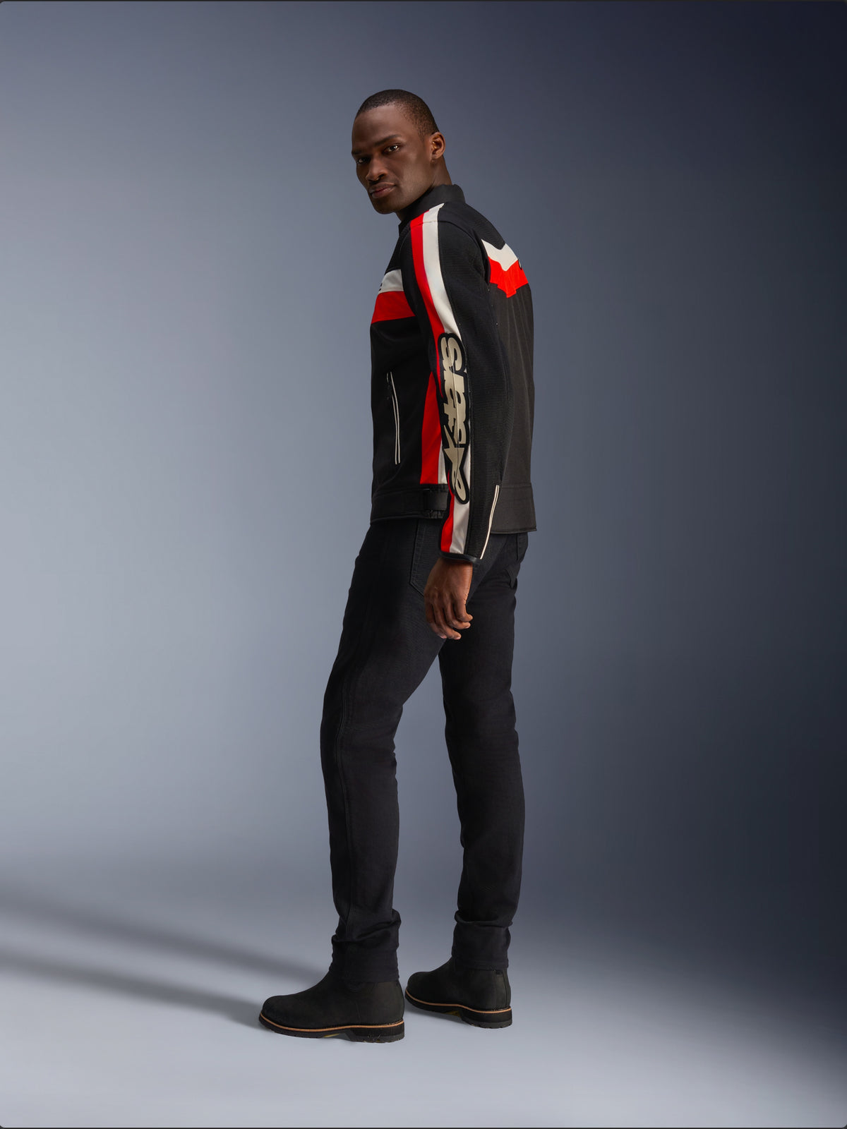 Ein Mann in der Alpinestars T-Dyno Air Jacke in Schwarz, Bright Red und Ecru, vintage-inspirierte Textil-Motorradjacke mit Mesh-Einsätzen, markantes Branding auf dem Ärmel, kombiniert mit schwarzer Hose und Stiefeln für einen modernen Biker-Look