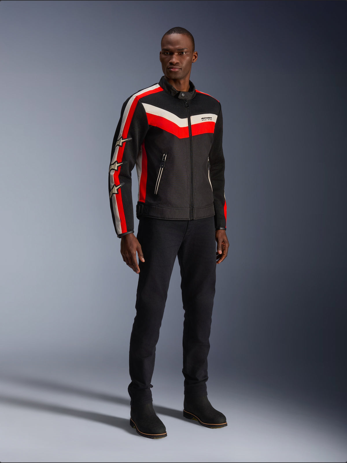Alpinestars T-Dyno Air Jacke, Schwarz, Hellrot und Ecru, Herren-Textilmotorradjacke mit 3D-Mesh-Einsätzen, roten und weißen Chevron-Streifen auf der Brust und Stern-Logos auf den Ärmeln, getragen von einem Model mit schwarzer Hose und Stiefeln.