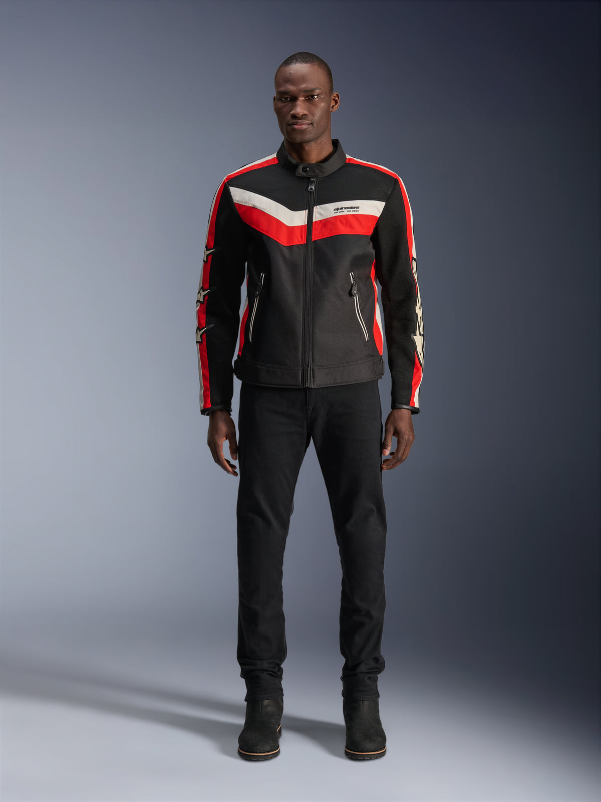 Alpinestars T-Dyno Air Jacke, Textil-Motorradjacke, Schwarz, Hellrot und Ecru, getragen von einem Model mit schwarzer Hose und Stiefeln, Vintage-inspiriertes Design mit markanten Farbstreifen auf der Brust, Stern-Logos an den Ärmeln und atmungsaktiver Mesh-Konstruktion für Fahrten in der Stadt.