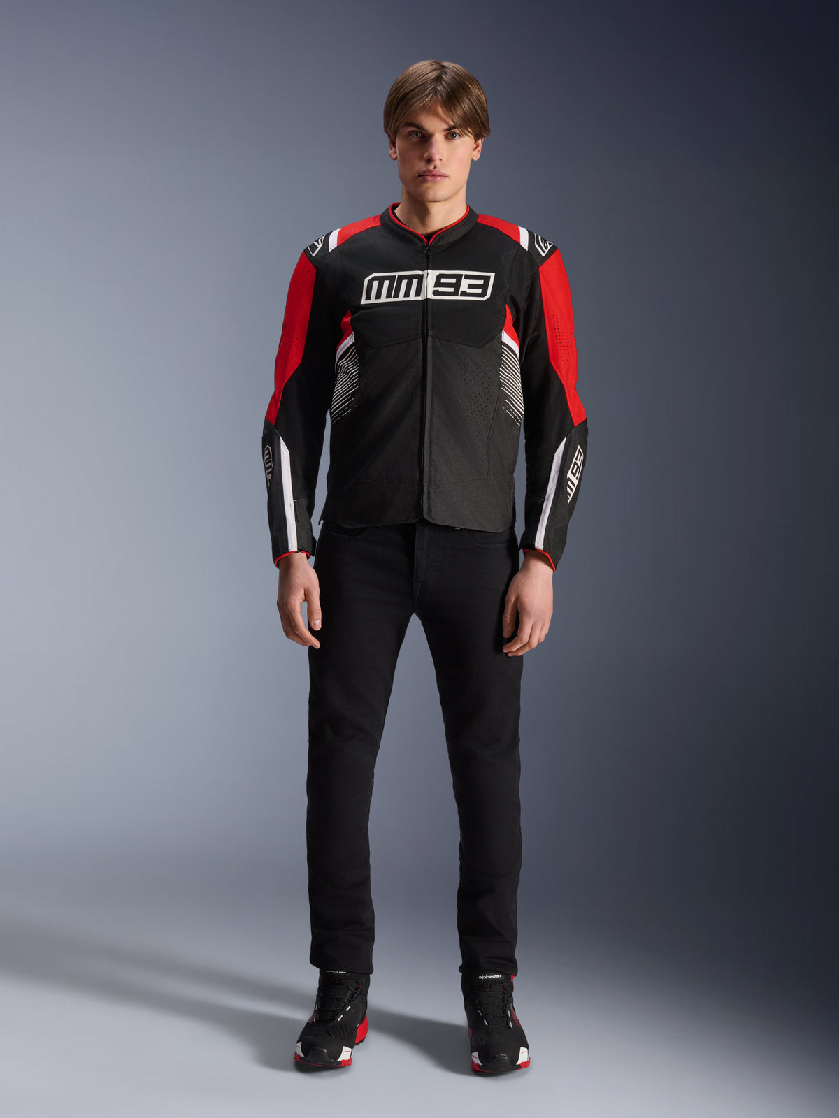 Männliches Model trägt die Alpinestars MM93 T-SPS Air V2 Jacke in Schwarz, leuchtendem Rot und Weiß, mit Marc Márquez MM93-Branding auf der Brust, Mesh-Einsätzen für Luftzirkulation, kombiniert mit einer schwarzen Hose und Sneakern in einem Studio-Setting.