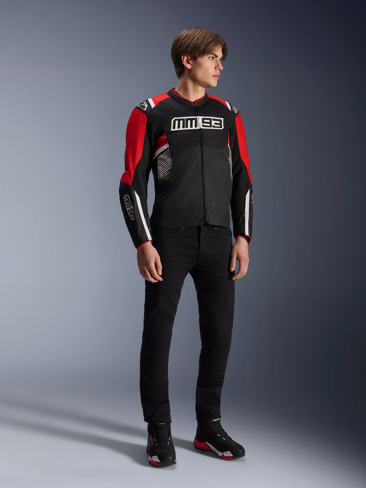 Männliches Model trägt die Alpinestars MM93 T-SPS Air v2 Jacke in Schwarz-Hellrot-Weiß, mit markantem MM93-Branding auf der Brust und den Ärmeln, kombiniert mit einer schwarzen Hose und Motorradstiefeln, sportlicher urbaner Motorradstil mit sichtbaren Mesh-Panels für optimale Belüftung.