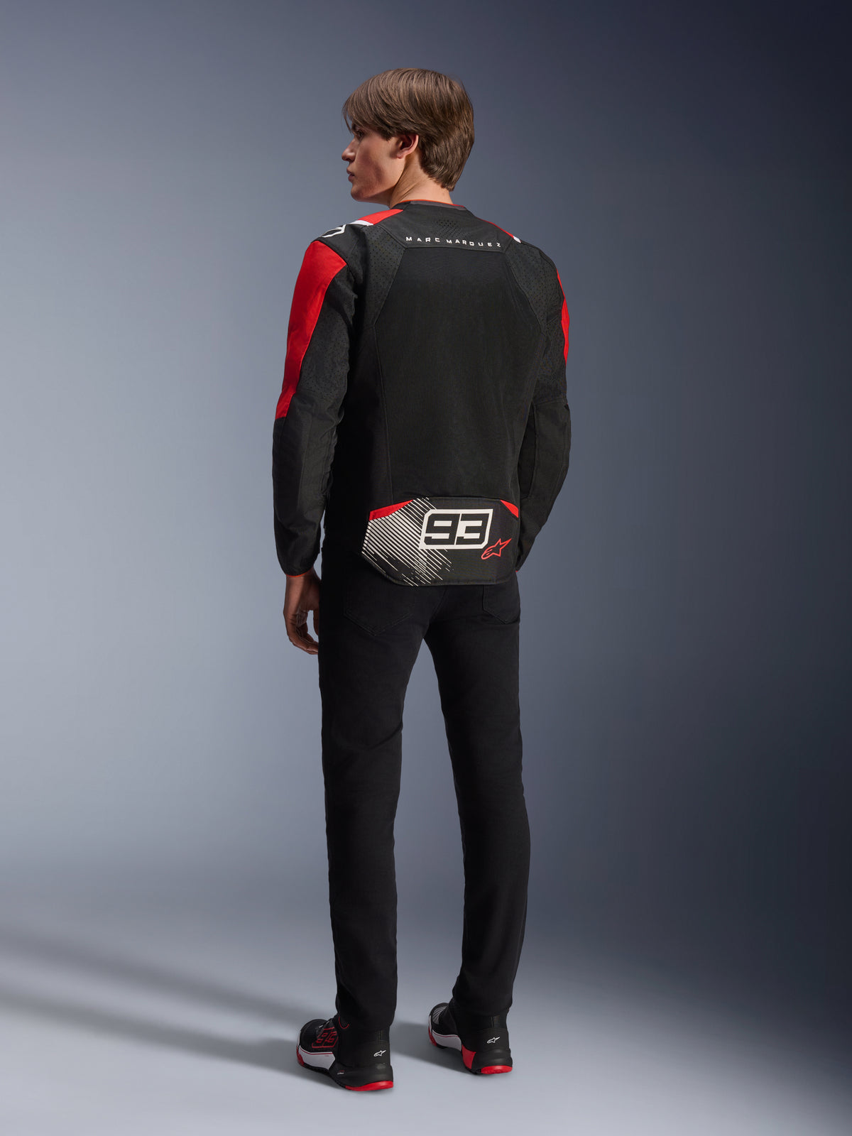 Modell von hinten in der Alpinestars MM93 T-SPS Air V2 Jacke, Schwarz mit leuchtend roten und weißen Details, mit 'MARC MARQUEZ' Schriftzug und großem '93' Logo auf der Rückseite, Mesh-Textilkonstruktion zur Belüftung, kombiniert mit einer schwarzen Hose und passenden schwarz-roten Motorradschuhen