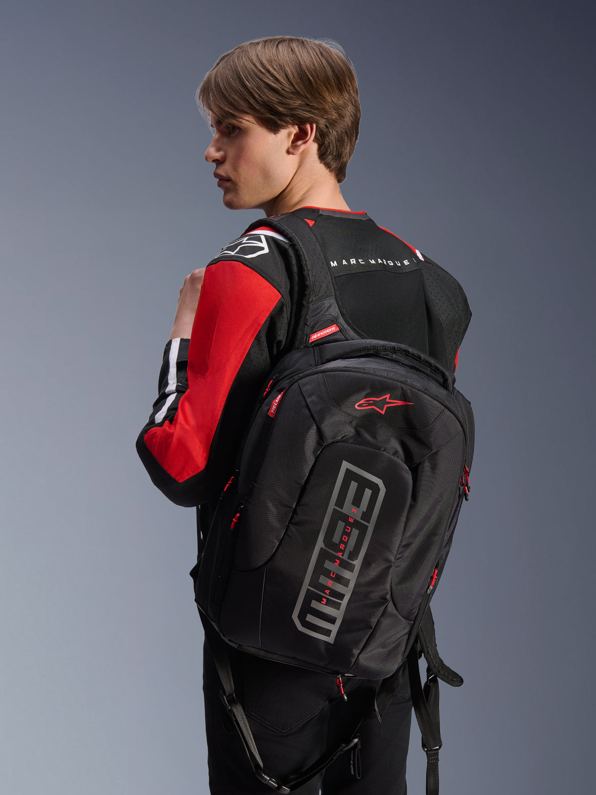Sac à dos Alpinestars MM93 City Hunter V2, noir, avec logo Astars rouge et marquage MM93 gris, porté par un motard avec un blouson de moto rouge et noir, équipement urbain de la collection Marc Márquez