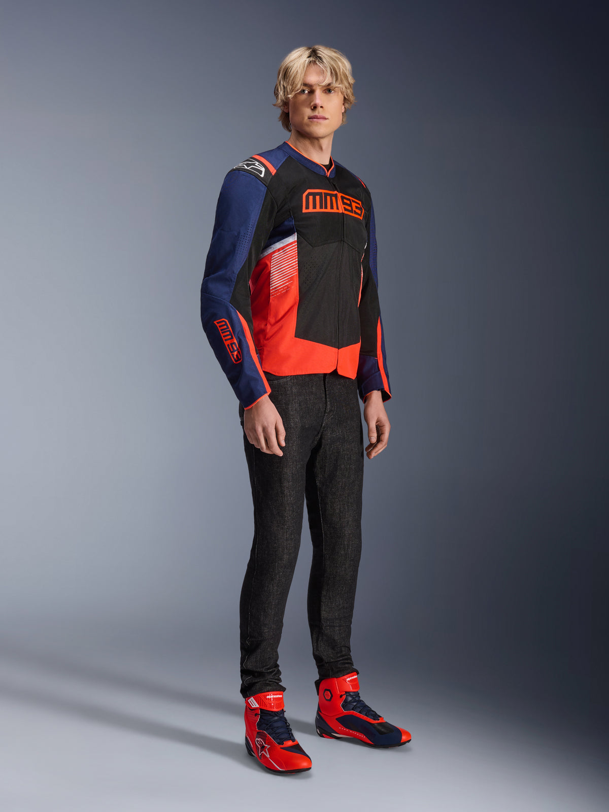 Männliches Model in der Alpinestars MM93 T-SPS Air V2 Jacke in Blue Navy, Schwarz und Rot Fluo, mit markantem MM93-Branding auf Brust und Ärmeln, kombiniert mit dunkler Jeans und passenden rot-blauen Motorradstiefeln, sportliche urbane Fahrausrüstung