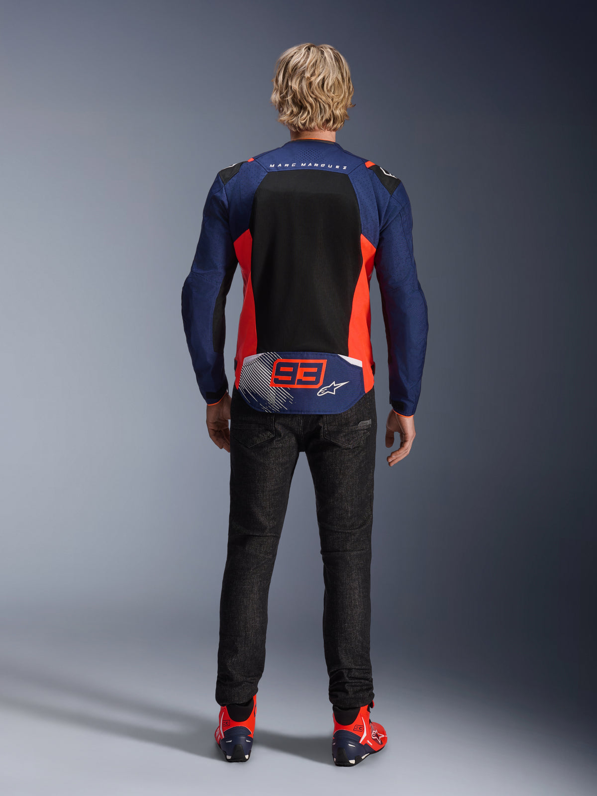 Rückansicht eines Modells mit der Alpinestars MM93 T-SPS Air V2 Jacke in Blue Navy mit Red Fluo und schwarzen Akzenten, großes Mesh-Panel am Rücken, '93' Marc Marquez Branding, kombiniert mit dunkler Denim-Hose und passenden Motorradstiefeln
