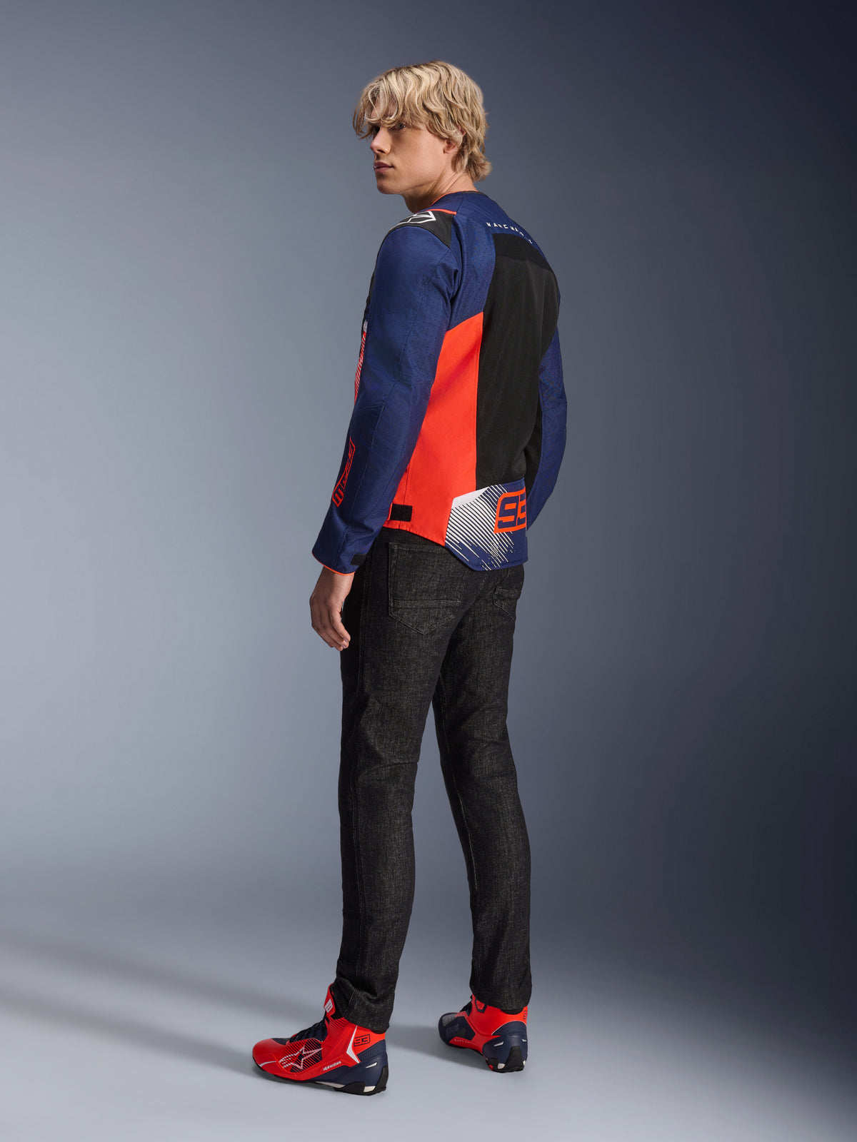 Alpinestars MM93 T-SPS Air V2 Jacke, Dunkelblau und Leuchtrot, von einem Modell von hinten getragen mit schwarzen Motorradjeans und passenden rot-blauen Schuhen, mit Mesh-Einsätzen und Marc Márquez 93 Branding.