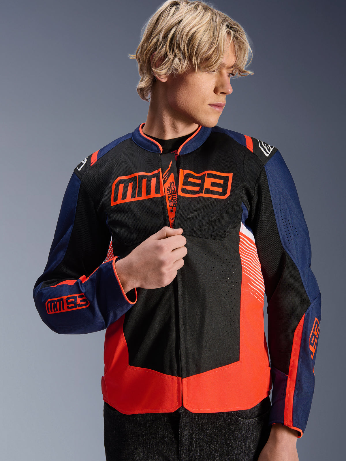 Alpinestars MM93 T-SPS Air V2 Jacke, Textil-Motorradjacke in Marineblau, Schwarz und Fluoreszierendem Rot, getragen von einem männlichen Model, das den Reißverschluss anpasst, mit markantem Marc Márquez MM93 Branding auf der Brust und den Ärmeln, belüftete Mesh-Einsätze für sommerliche Straßenfahrten.