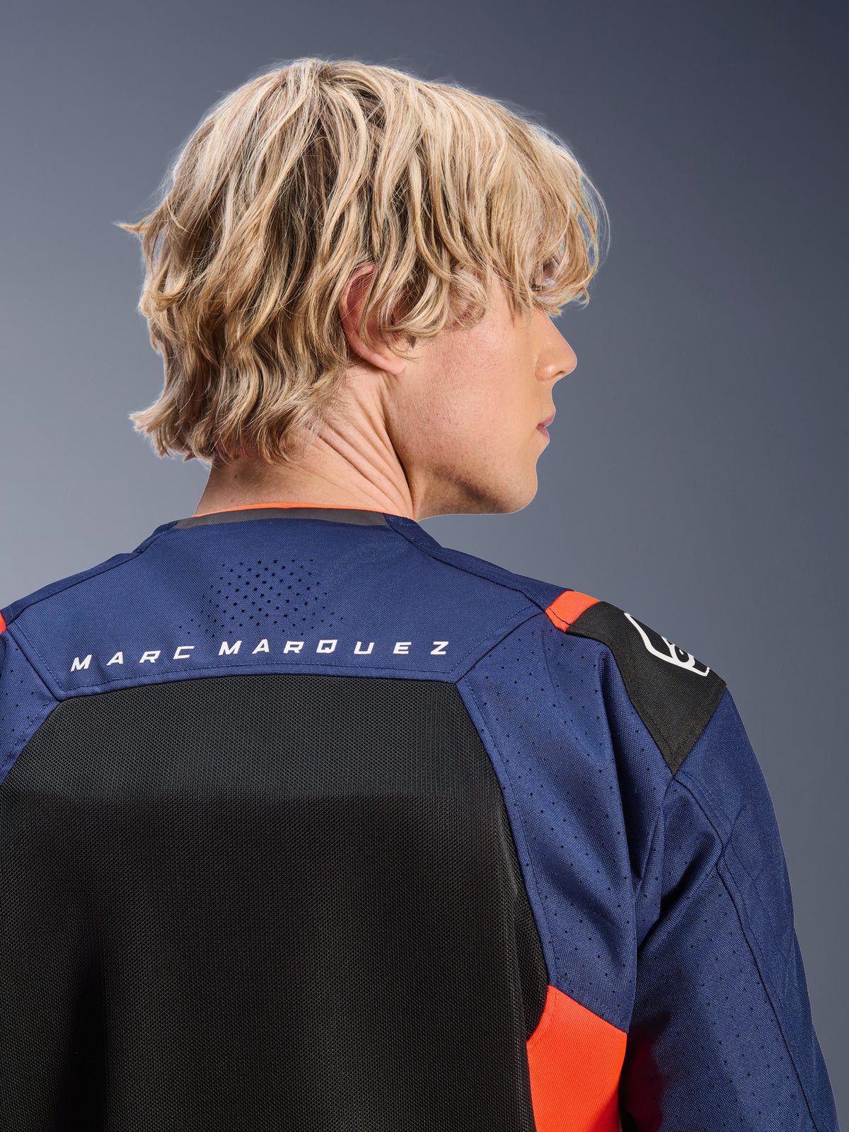 Alpinestars MM93 T-SPS Air V2 Jacke, Navy-Blau und Schwarz mit fluoreszierend roten Akzenten, Nahaufnahme des oberen Rückens mit „MARC MARQUEZ“-Branding, atmungsaktive Mesh-Panels und perforierte Textilkonstruktion, getragen von einem blonden Model.