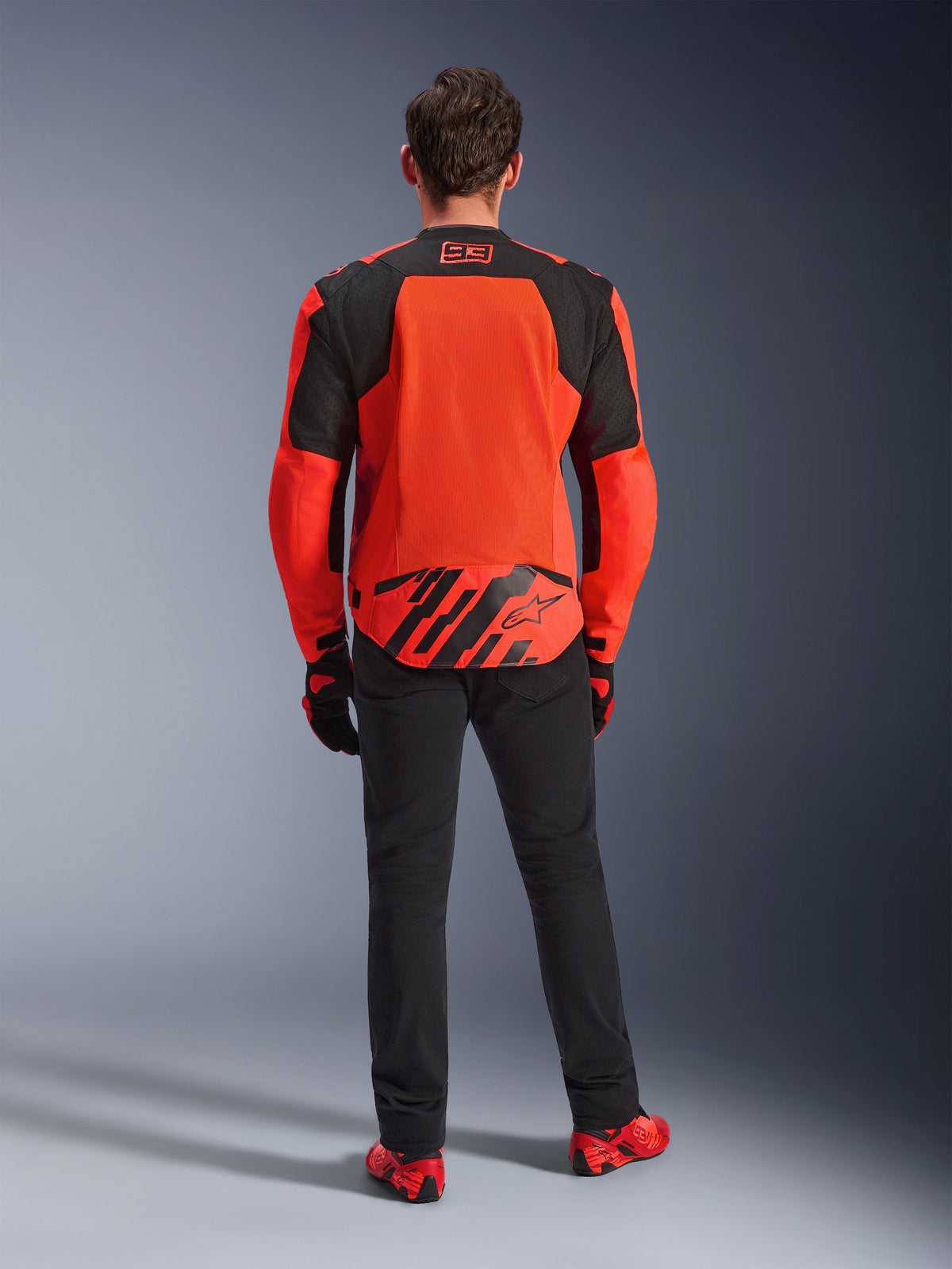Vue arrière de la veste MM93 T-SPS Air V2, rouge fluo et noir, veste de moto en textile avec de grands panneaux en mesh dans le dos pour la ventilation, portée avec un pantalon noir et des chaussures rouges, arborant le marquage Marc Marquez 93