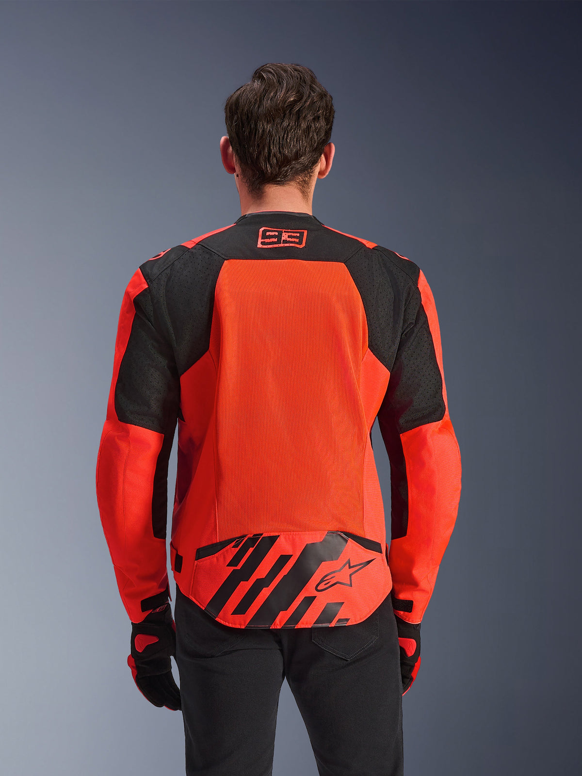 Veste Alpinestars MM93 T-SPS Air V2, vue de dos, rouge fluo et noir, avec le logo de course 93 sur le col, un grand panneau en maille dans le dos pour la ventilation, un rabat au bas du dos avec le logo étoile Alpinestars et des rayures géométriques, portée avec un pantalon noir et des gants de moto