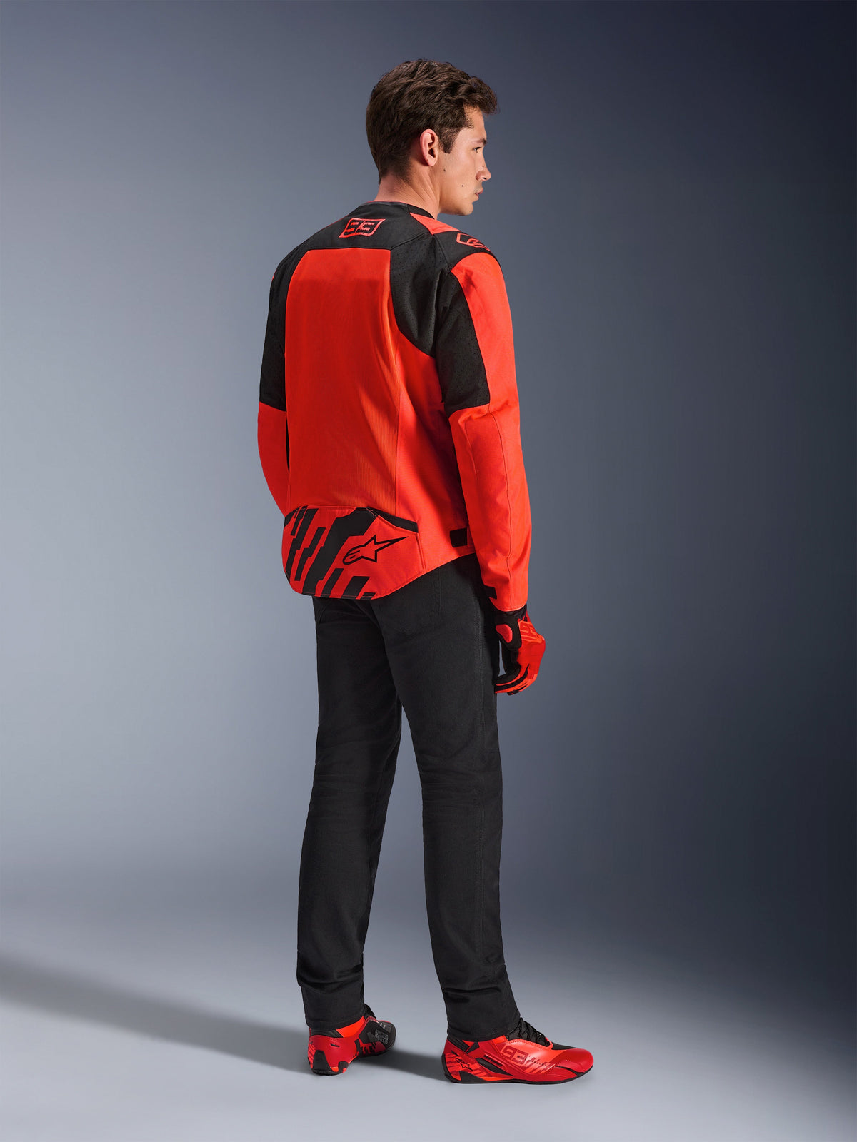 Veste Alpinestars MM93 T-SPS Air V2 en rouge fluo et noir, vue de trois-quarts arrière sur un mannequin, avec marquage Marc Marquez 93 sur le col et le bas du dos, portée avec un pantalon noir et des chaussures de moto rouges.