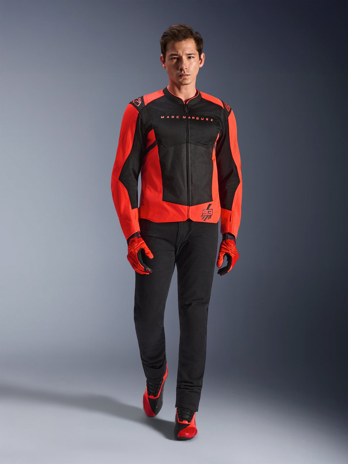 Veste Alpinestars MM93 T-SPS Air V2, rouge fluo et noir, portée par un mannequin avec des gants rouges assortis et un pantalon noir, arborant le marquage Marc Marquez et le logo 93, veste de moto de sport ventilée pour la conduite urbaine