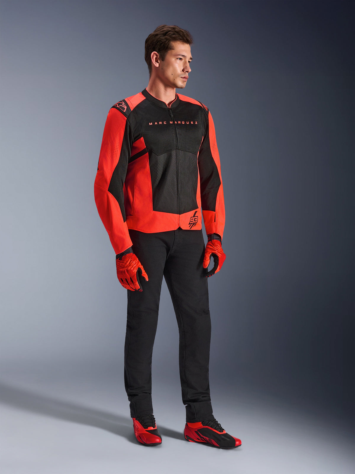 Veste Alpinestars MM93 T-SPS Air V2, rouge fluo et noir, portée par un mannequin avec un pantalon noir assorti, des gants rouges et noirs et des chaussures de moto rouges, avec marquage Marc Marquez et logo 93, équipement de conduite urbaine au style sportif