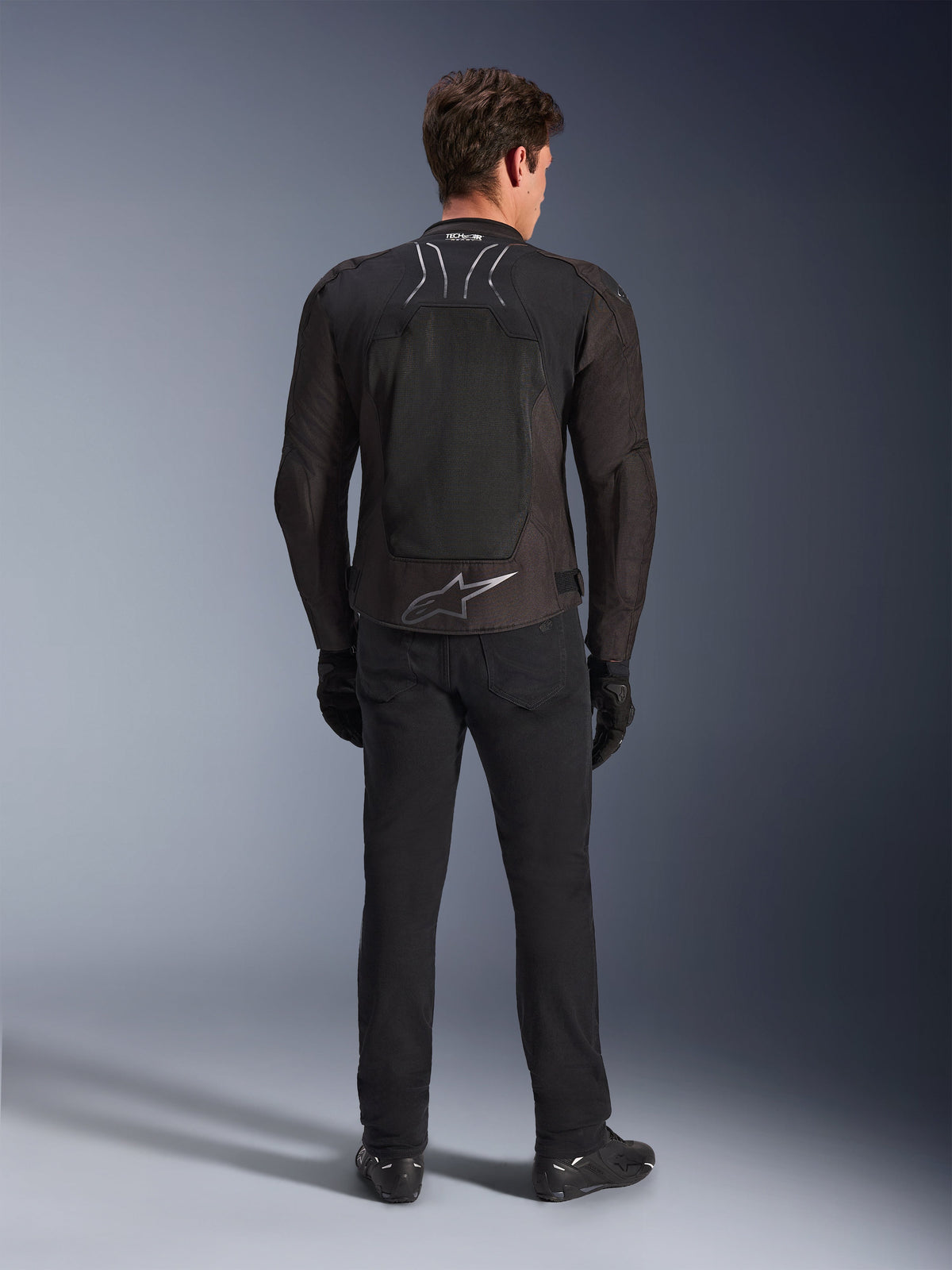 Alpinestars SMX Air Jacket, Black Anthracite, achteraanzicht op mannelijk model, met een groot zilveren Alpinestars-logo op de onderrug en high-flow mesh-panelen, gedragen met zwarte motorbroek, handschoenen en laarzen, sportieve motoruitrusting