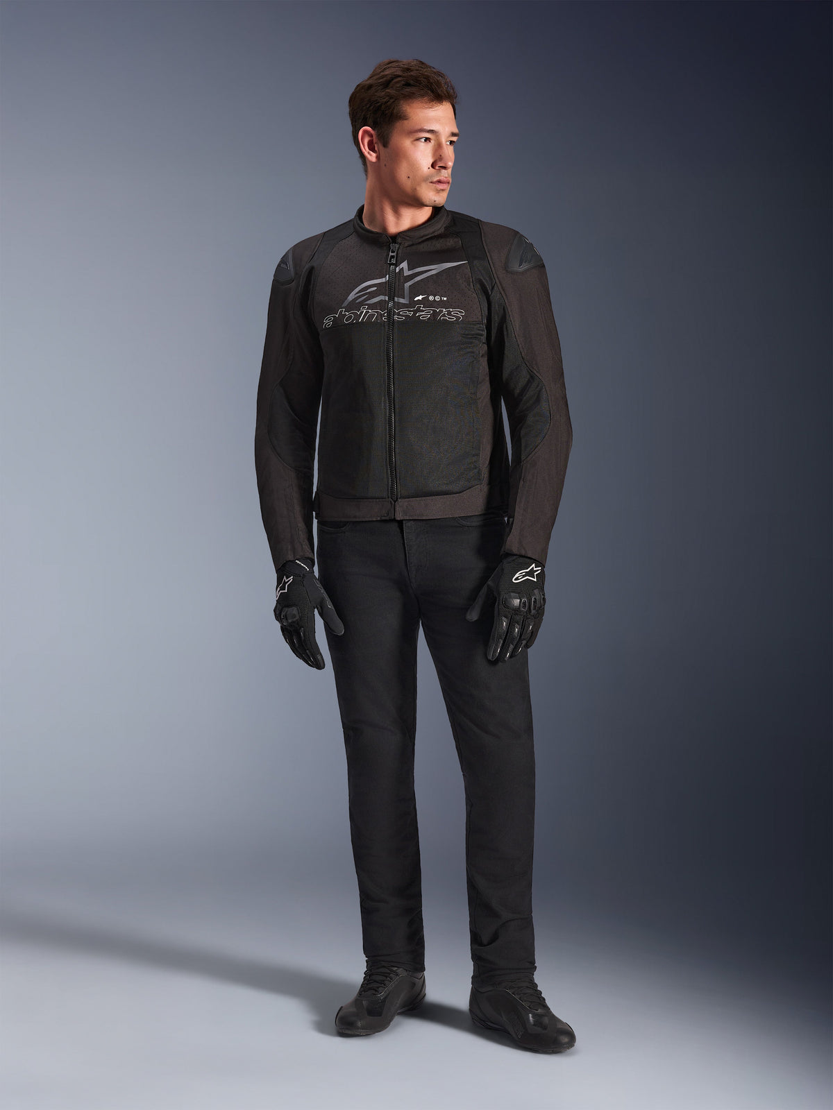 Mannelijk model draagt de Alpinestars SMX Air Jacket, een Black Anthracite motorjas met geventileerde mesh-panelen en een grijs logo op de borst, gecombineerd met een zwarte broek en beschermende handschoenen, studio-opname met een gradiënt achtergrond