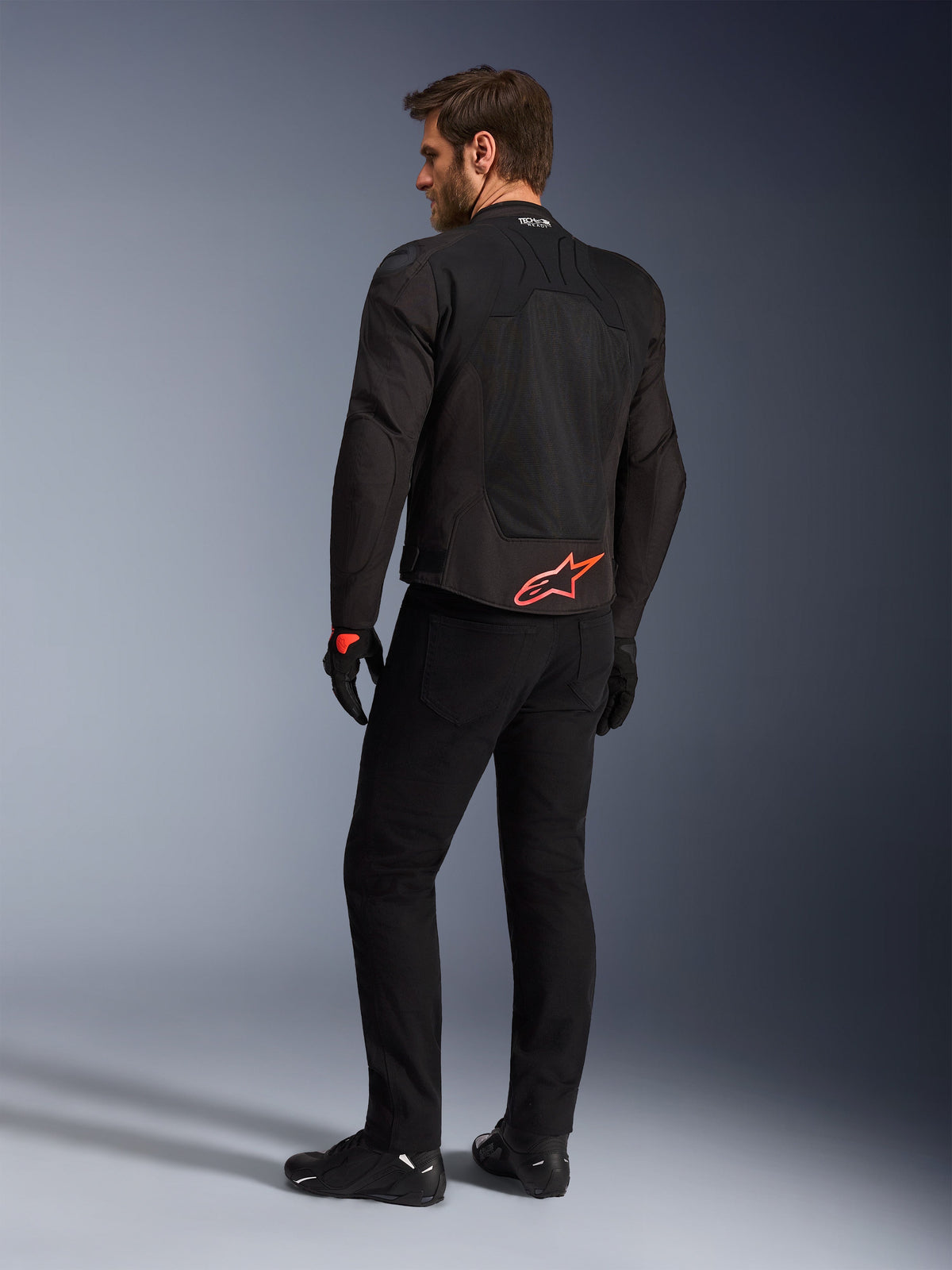 Vue arrière d'un mannequin portant la veste Alpinestars SMX Air en noir, arborant un logo Alpinestars rouge vif en bas du dos, le marquage « Tech-Air Ready » sur le col et des panneaux en maille technique pour la ventilation, associée à un pantalon et des gants de moto noirs.