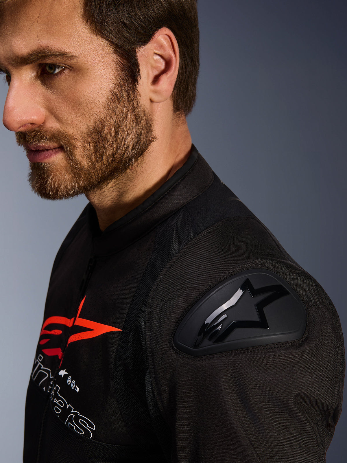 Veste Alpinestars SMX Air, noir et rouge, gros plan sur la protection d'épaule DFS Lite avec logo en étoile, portée par un homme barbu, veste de moto textile sport et racing.