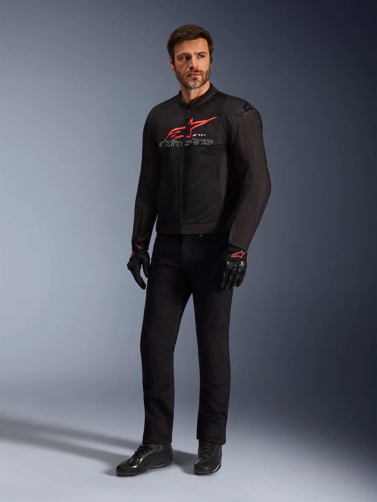 Modèle masculin portant la veste Alpinestars SMX Air en noir, rouge vif et blanc, avec un pantalon technique noir, des gants noirs et rouges et des chaussures de moto noires, logo rouge distinctif sur la poitrine, photo de studio en pied pour équipement de moto sportive
