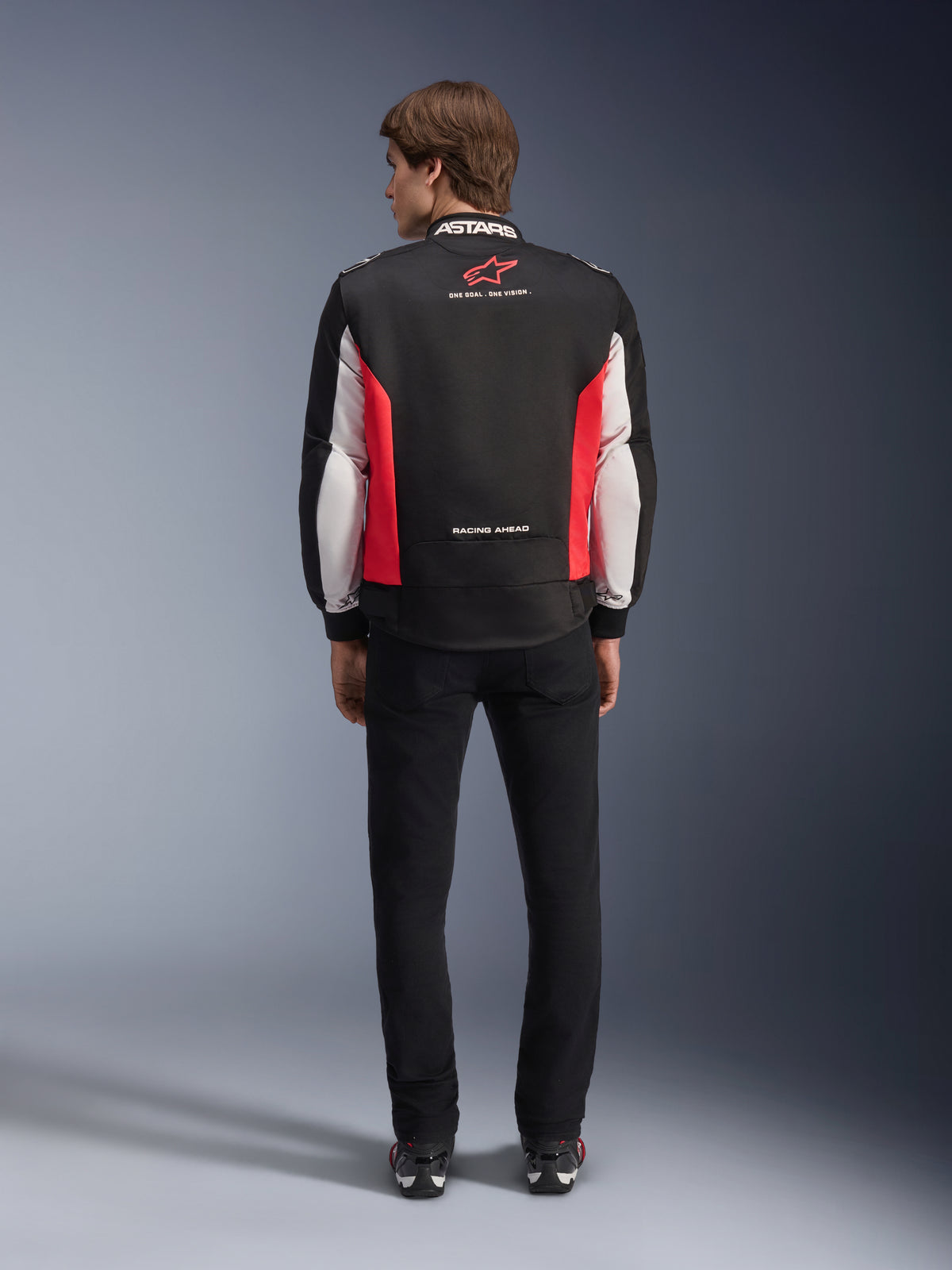 Veste Alpinestars Monza Sport, veste de moto en textile noir, rouge vif et blanc, vue arrière sur un mannequin, avec logo étoile rouge et marquage « ASTARS » sur le col avec slogans, portée avec un pantalon noir et des bottes de moto