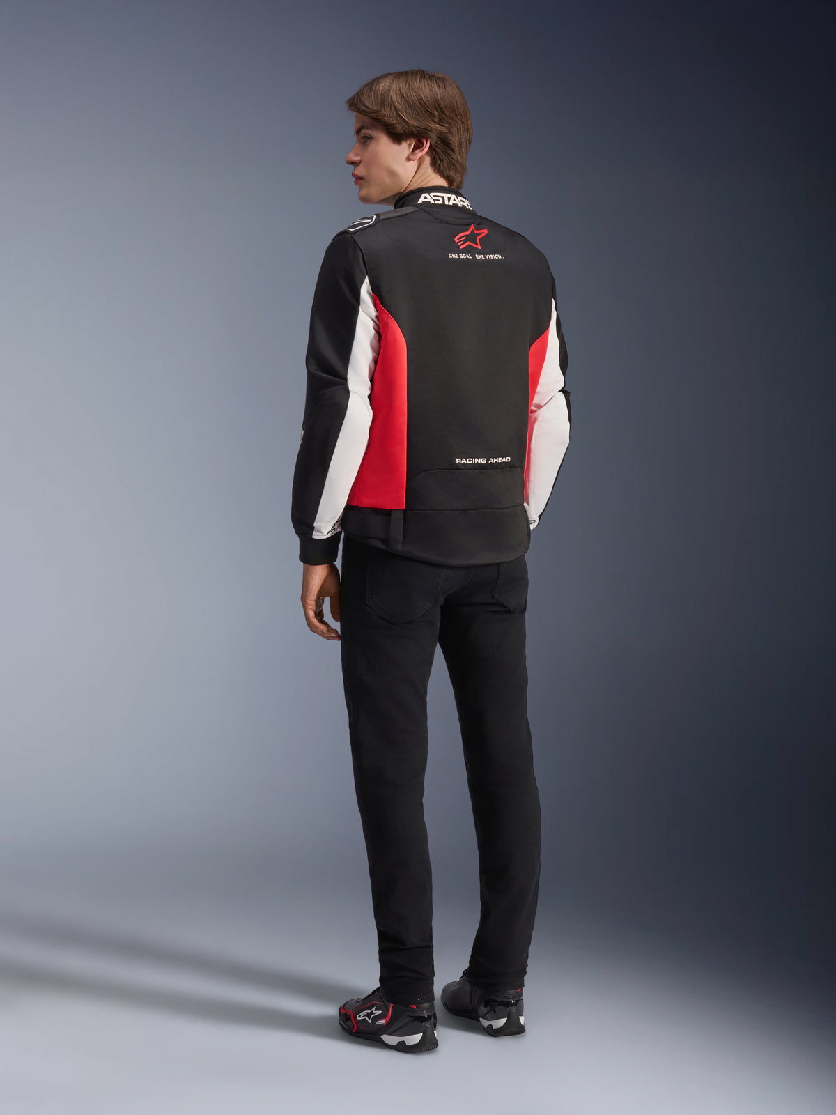 Veste Alpinestars Monza Sport, veste de moto en textile, noir, rouge vif et blanc, vue de dos du mannequin présentant les panneaux latéraux rouges et les détails blancs sur les manches, logo Alpinestars et texte « ONE GOAL. ONE VISION. », associée à un pantalon noir et des chaussures de moto pour un look d'inspiration sportive