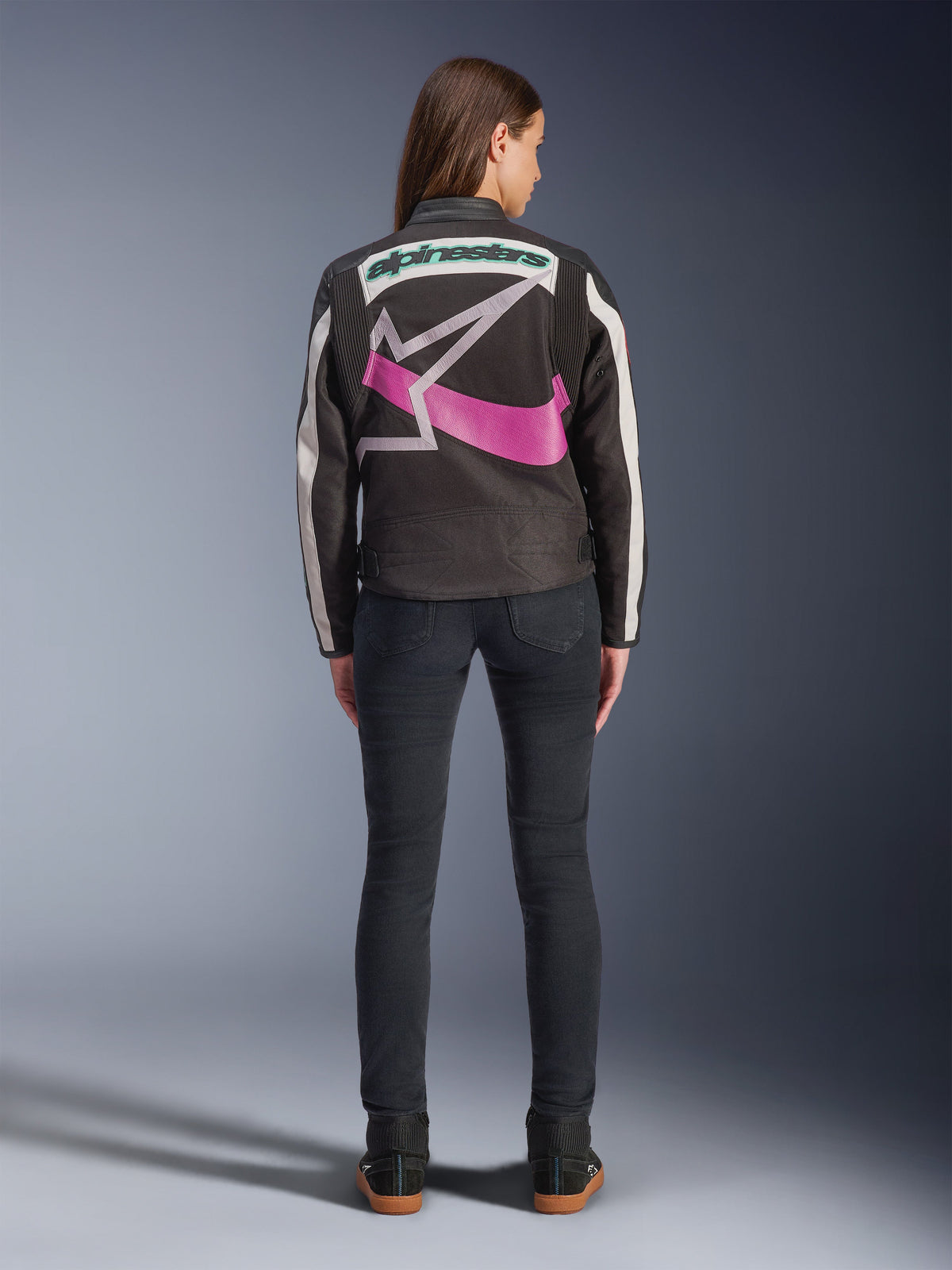 Rückansicht einer Frau in der Alpinestars Idles Jacke, Schwarz mit cremeweißen Streifen, mit einem großen lila-weißen Alpinestars Stern-Logo und grünem Schriftzug auf dem Rücken, kombiniert mit einer dunklen Slim-Fit-Jeans, vintage-inspirierte urbane Motorradbekleidung