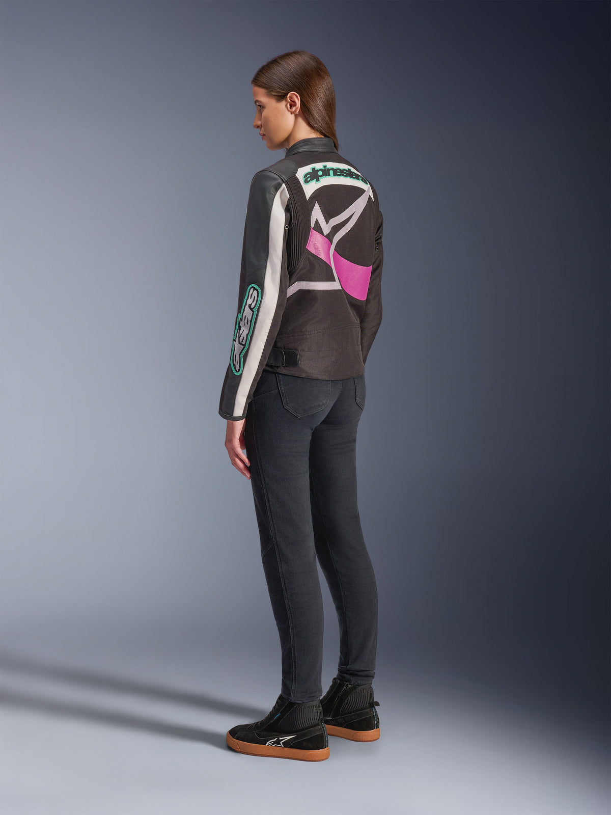 Alpinestars Idles Damenjacke, Motorradjacke, Schwarz, Off-White und Lila, Rückansicht am Modell, mit großem umlaufendem Alpinestars-Logo und von Vintage-Rennsport inspiriertem Design, kombiniert mit dunklen Jeans und schwarzen Schuhen.