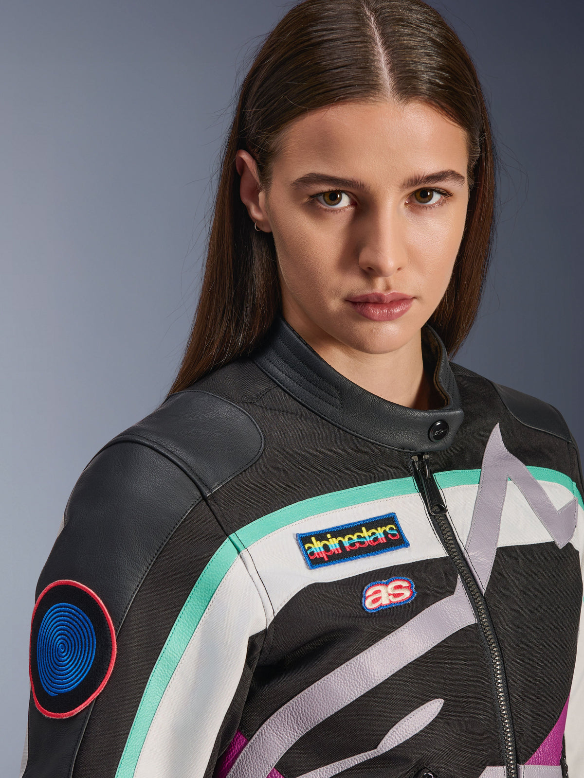Alpinestars Idles Damenjacke, Motorradjacke, Schwarz, Off-White und Lila mit blaugrünen Akzenten, Nahaufnahme mit farbenfrohen Alpinestars Heritage-Aufnähern und Schulterdetails aus Ziegenleder, Vintage-inspiriertes Design für urbanes Fahren.