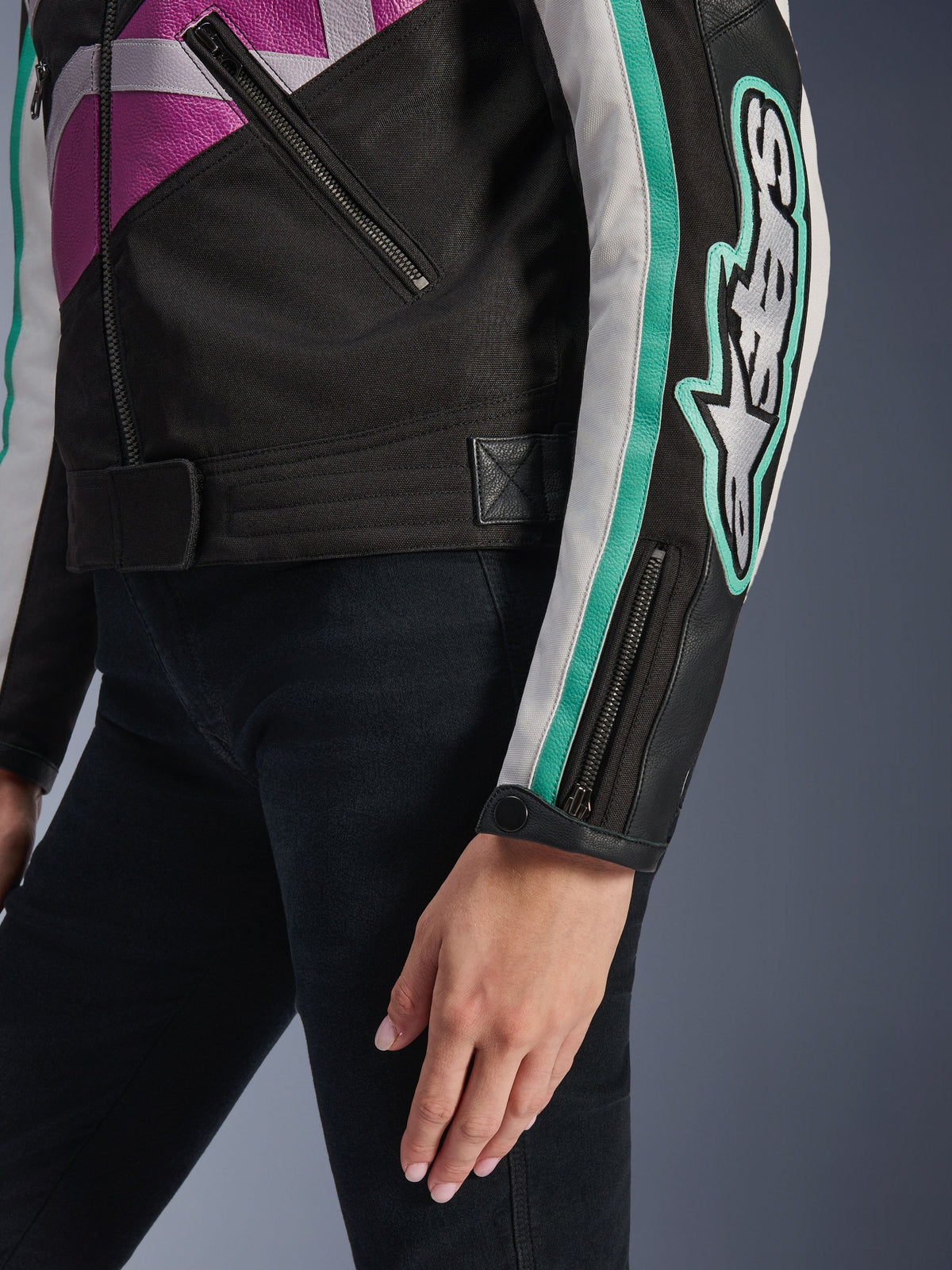 Alpinestars Idles Damenjacke, Nahaufnahme von Ärmel und seitlichem Torso, Schwarz mit lila, weißen und türkisfarbenen Akzenten, mit auffälligem Alpinestars-Logo am Arm, Ärmelbündchen mit Reißverschluss und Druckknopf sowie Taillenverstellriemen, vintage-inspirierte Motorradbekleidung.